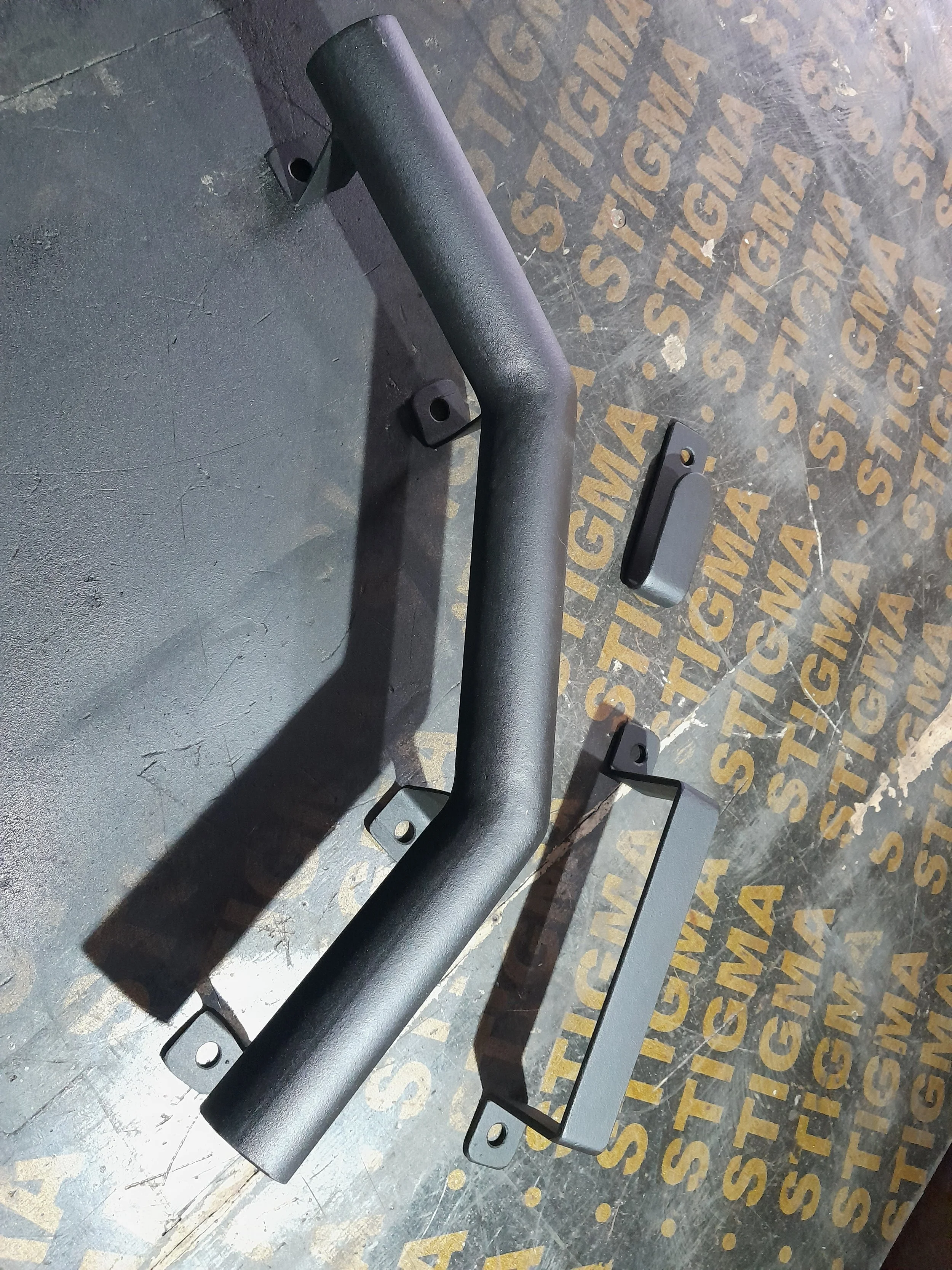 Black metal handlebars and brackets on a textured surface with "STIGMA" printed in gold.