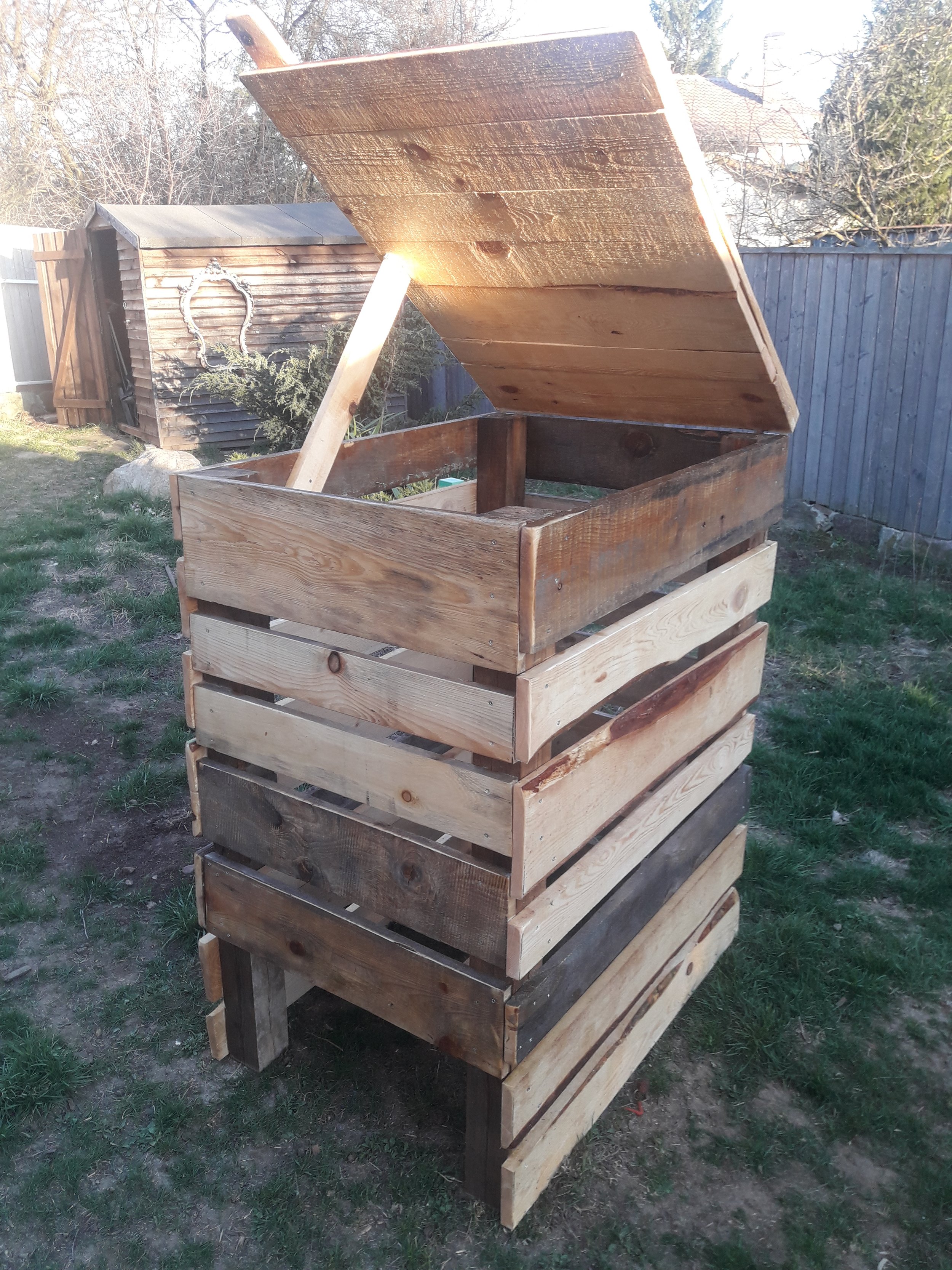 Wooden compost bin with an open lid in a backyard setting, surrounded by grass and trees.
