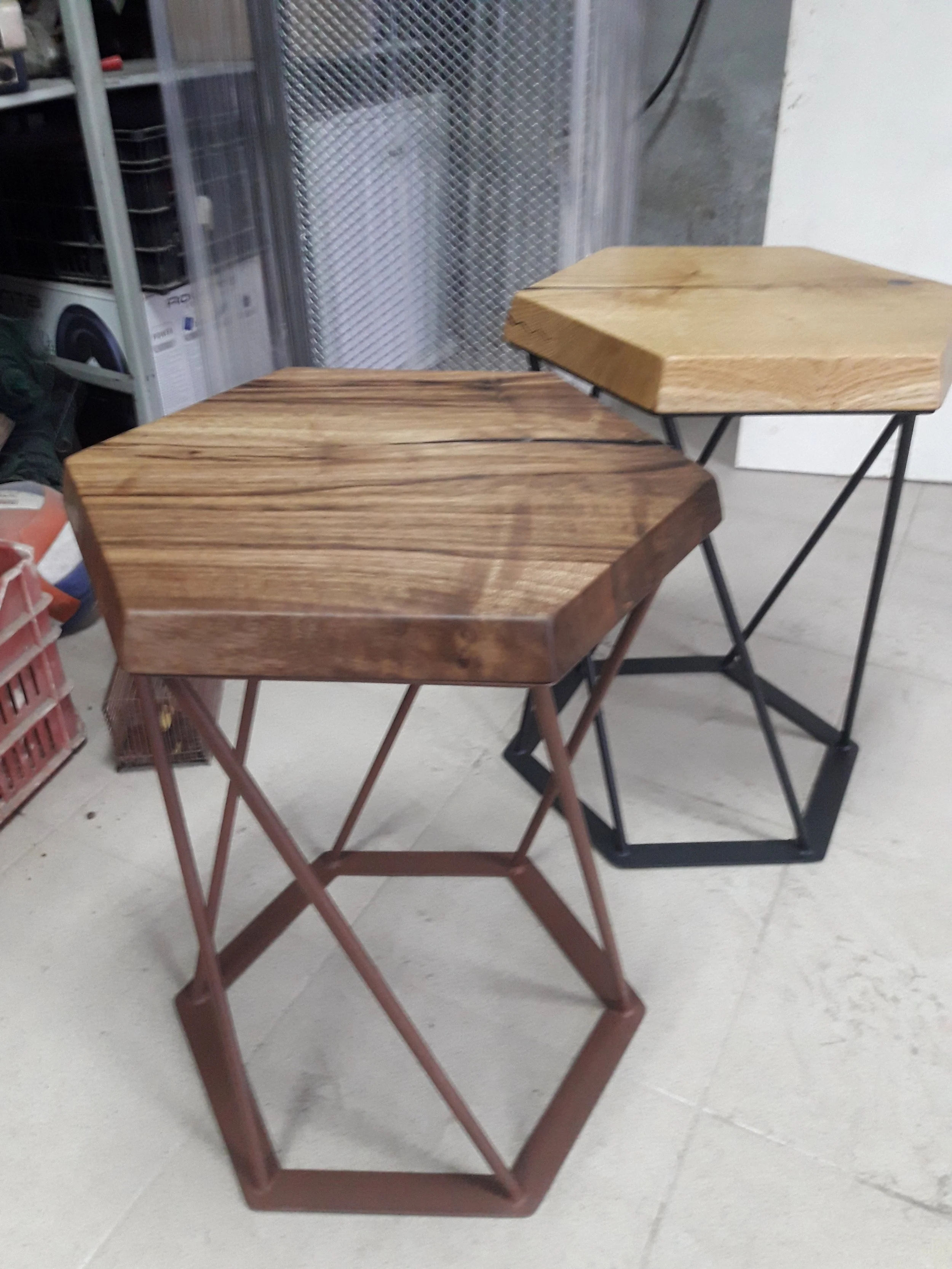 Two hexagonal wooden tables with metal bases, one with a darker wood top and rust-colored legs, the other with a lighter wood top and black legs, set on a tiled floor.