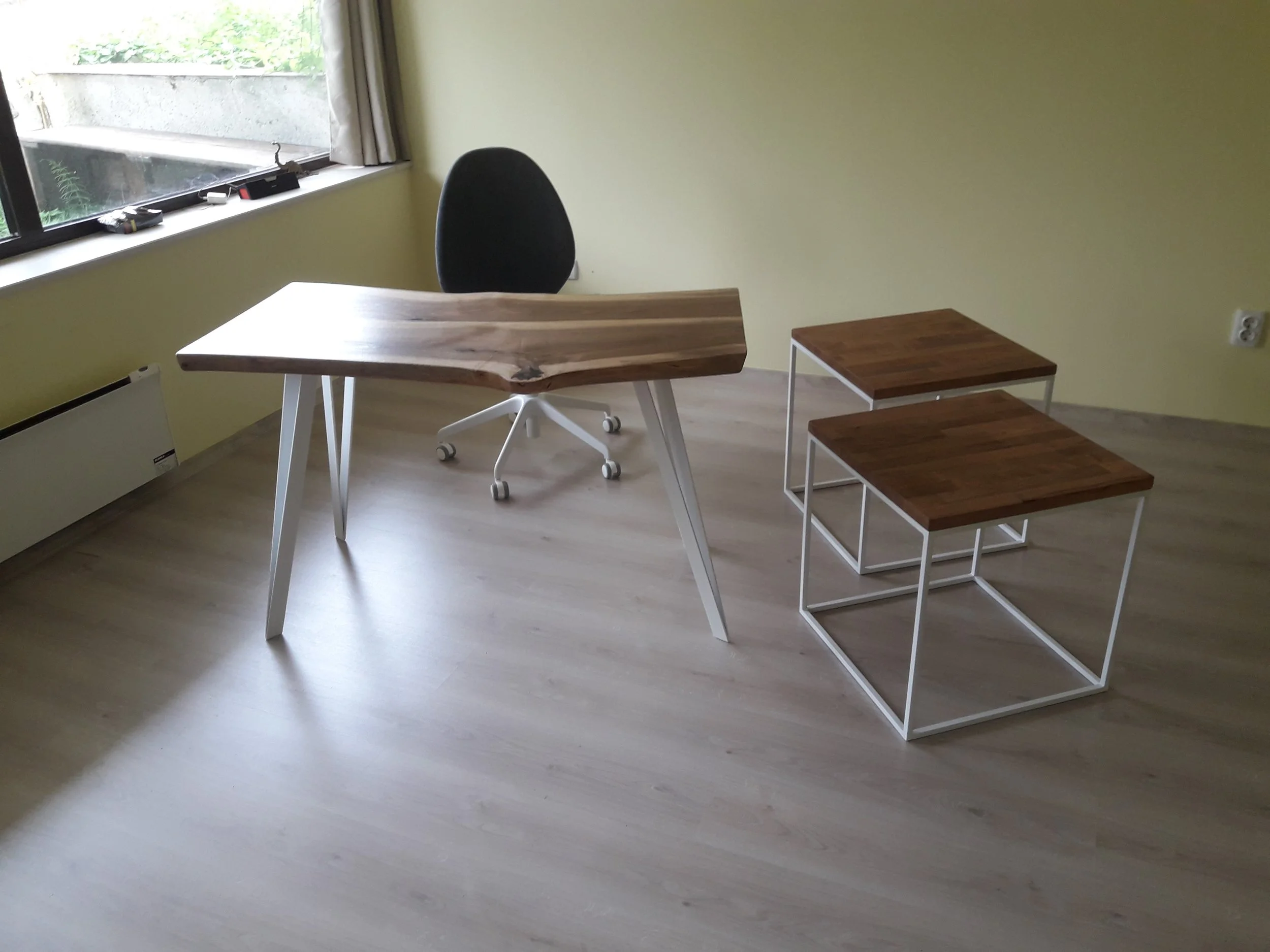 Modern office with a unique wooden desk, two square wooden stools with metal frames, and an office chair. The room has a large window with natural light and a beige wall.