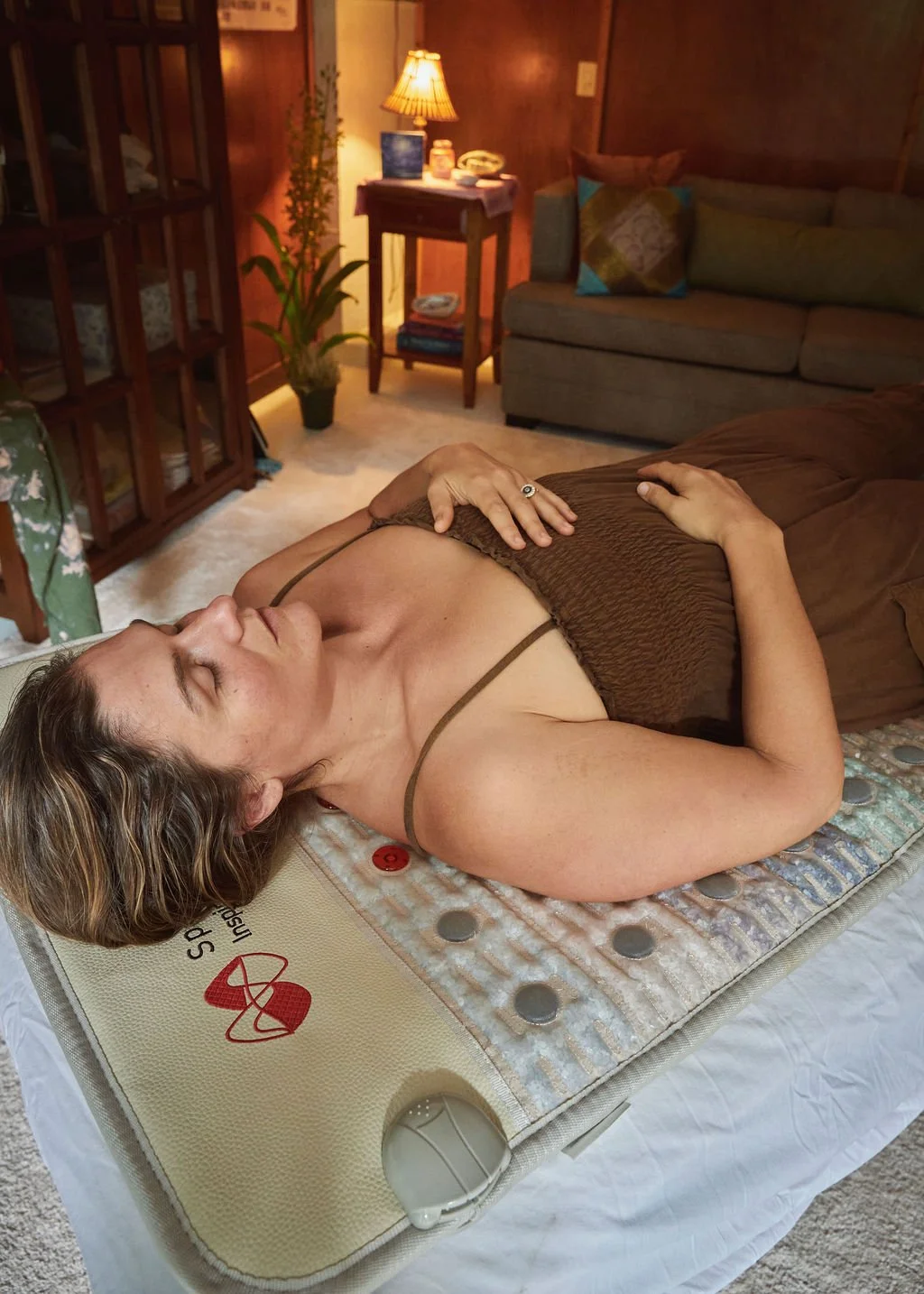 A group of people practicing yoga indoors, lying on mats with bolster pillows, overlooking a lush green outdoor view with trees and a waterfall.