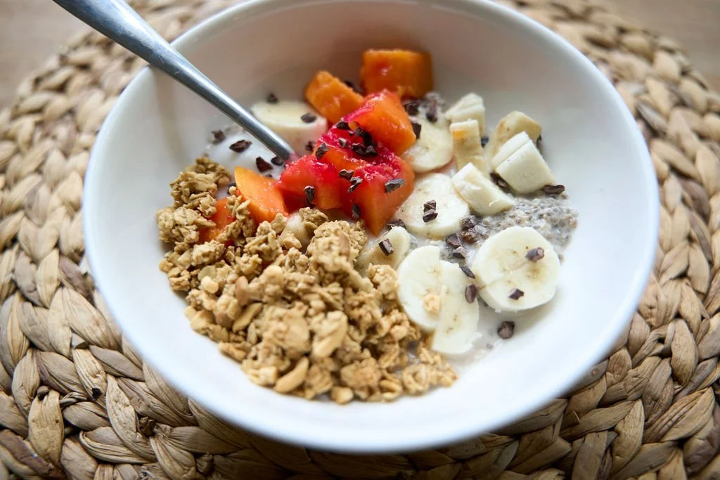 A breakfast table featuring a colorful patterned tablecloth. On the table, there's a plate with sliced bananas and raisins, a bowl with sliced papaya and small red peppers, a dish with grapes, a plate of chopped oranges, a bowl of cereal, a French press, a jar with a lid, a cup of coffee, and empty plates and silverware.