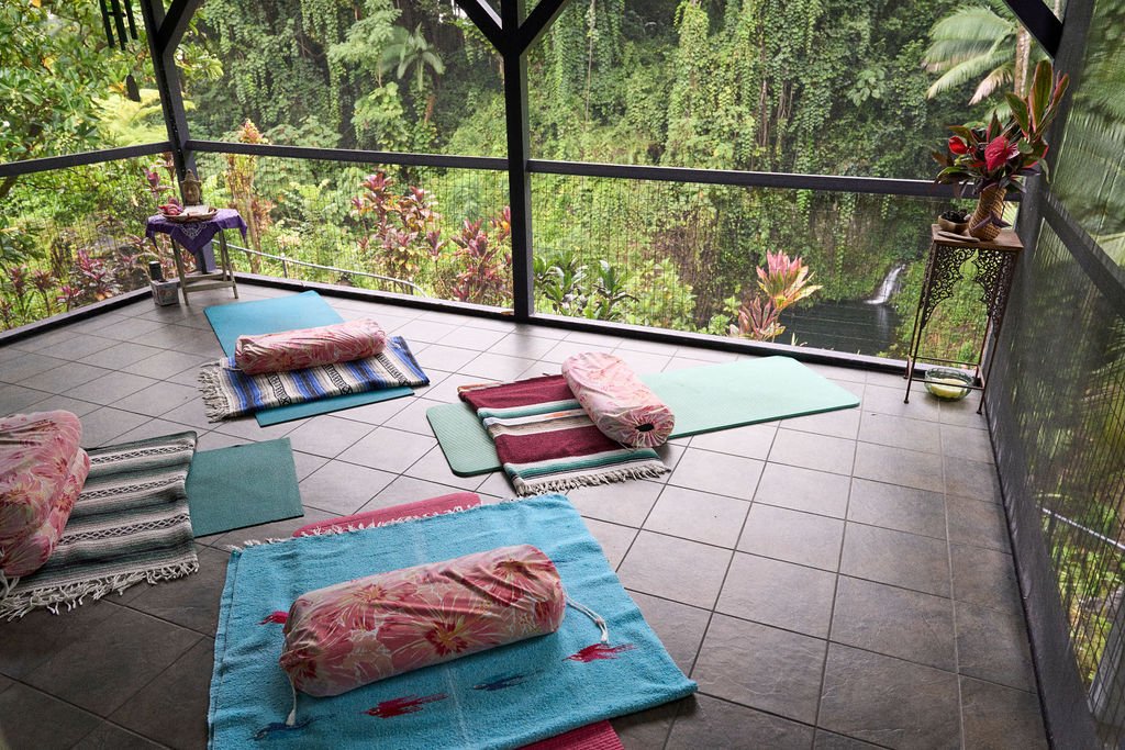 Person lying on grassy area by the ocean, surrounded by palm trees and rocks, wearing a black outfit. The sky is cloudy.