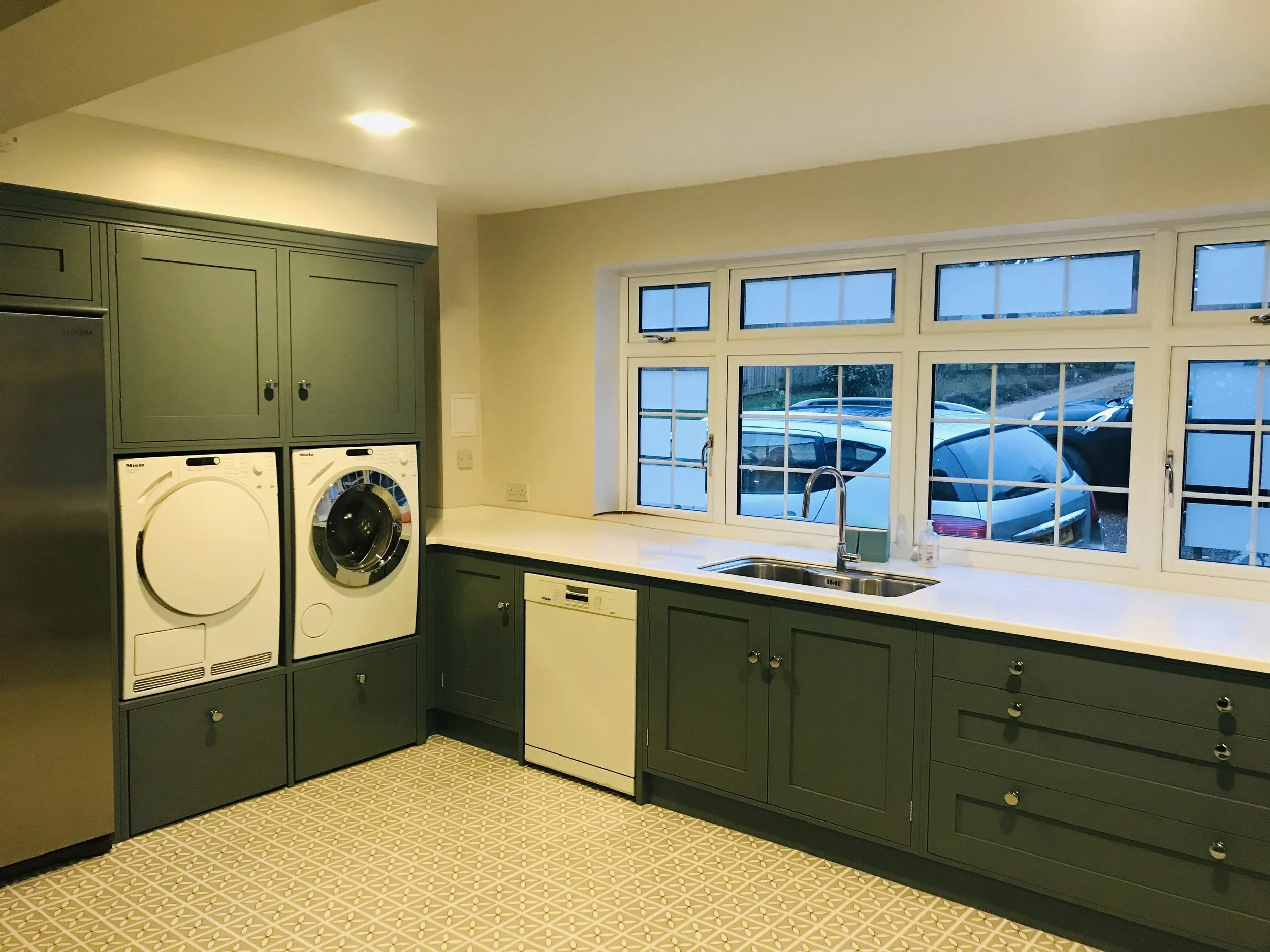 Laundry room with green cabinets, white countertop, washer, dryer, dishwasher, window with parking lot outside.