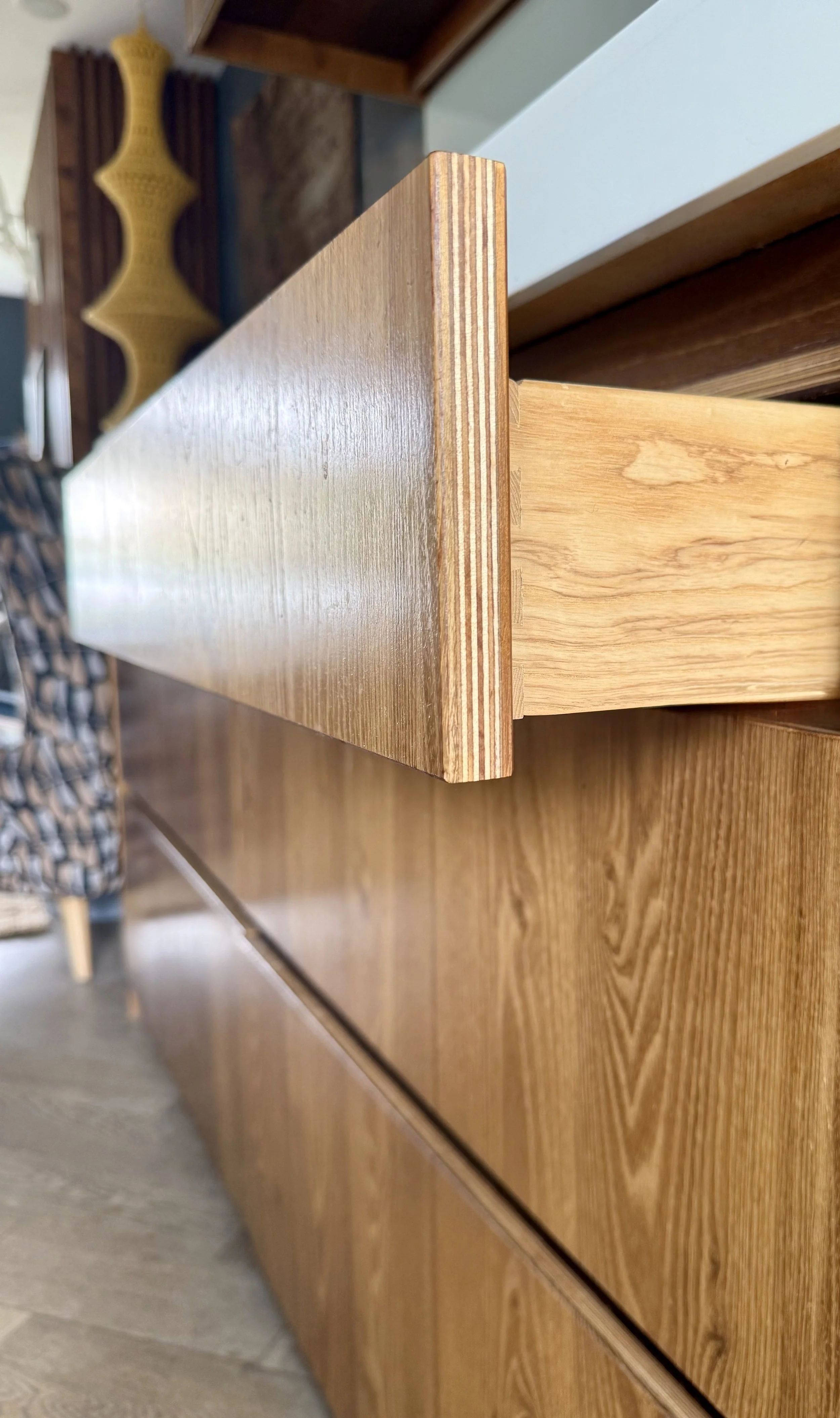 Close-up view of a wooden drawer slightly pulled out from a matching wooden cabinet, showing the wood grain and dovetail joints on the side. In the background, a yellow decorative object is visible.