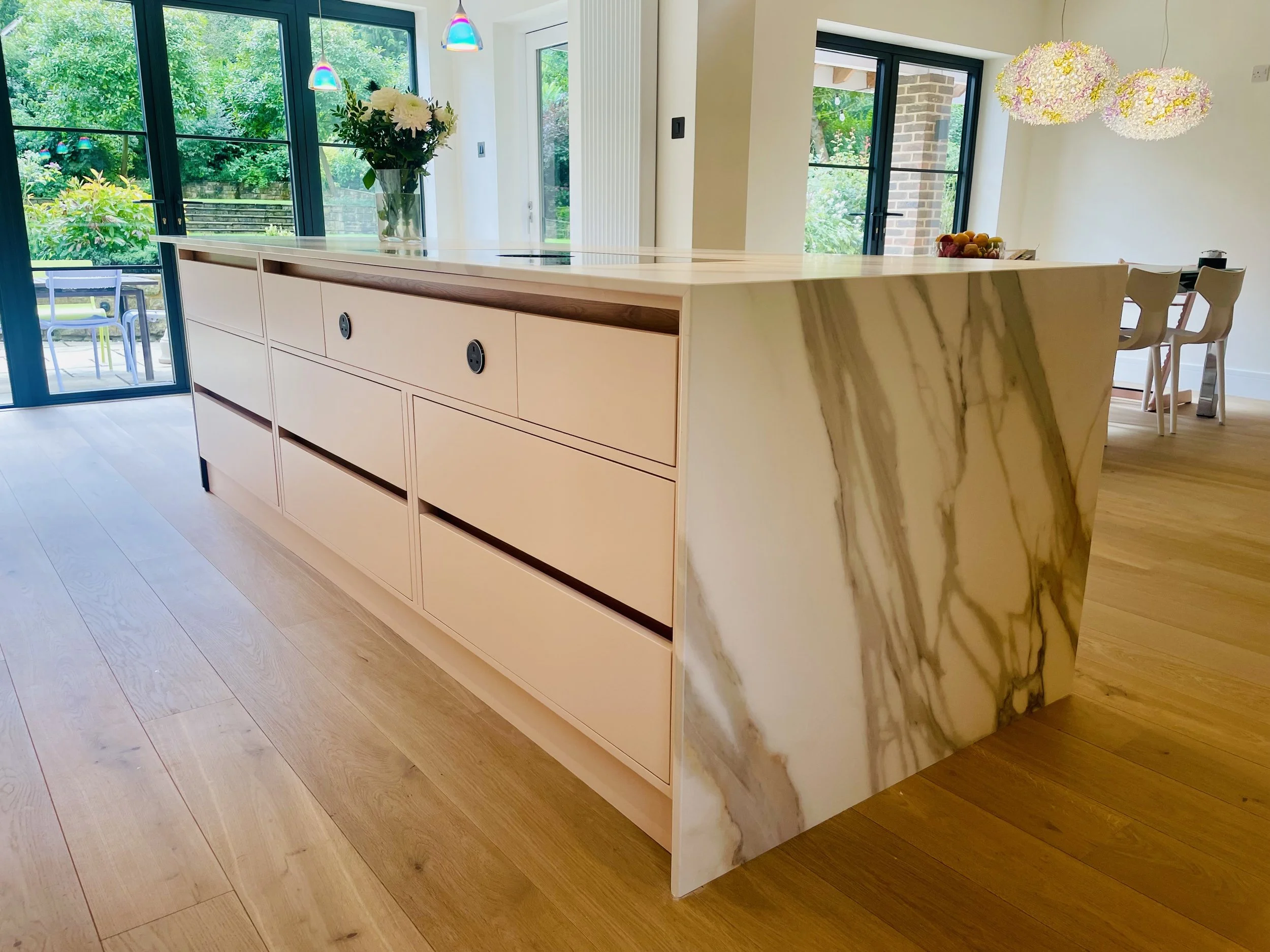 Modern kitchen island with marble side panel and light wood drawers in a bright open-plan room with large windows and dining area.