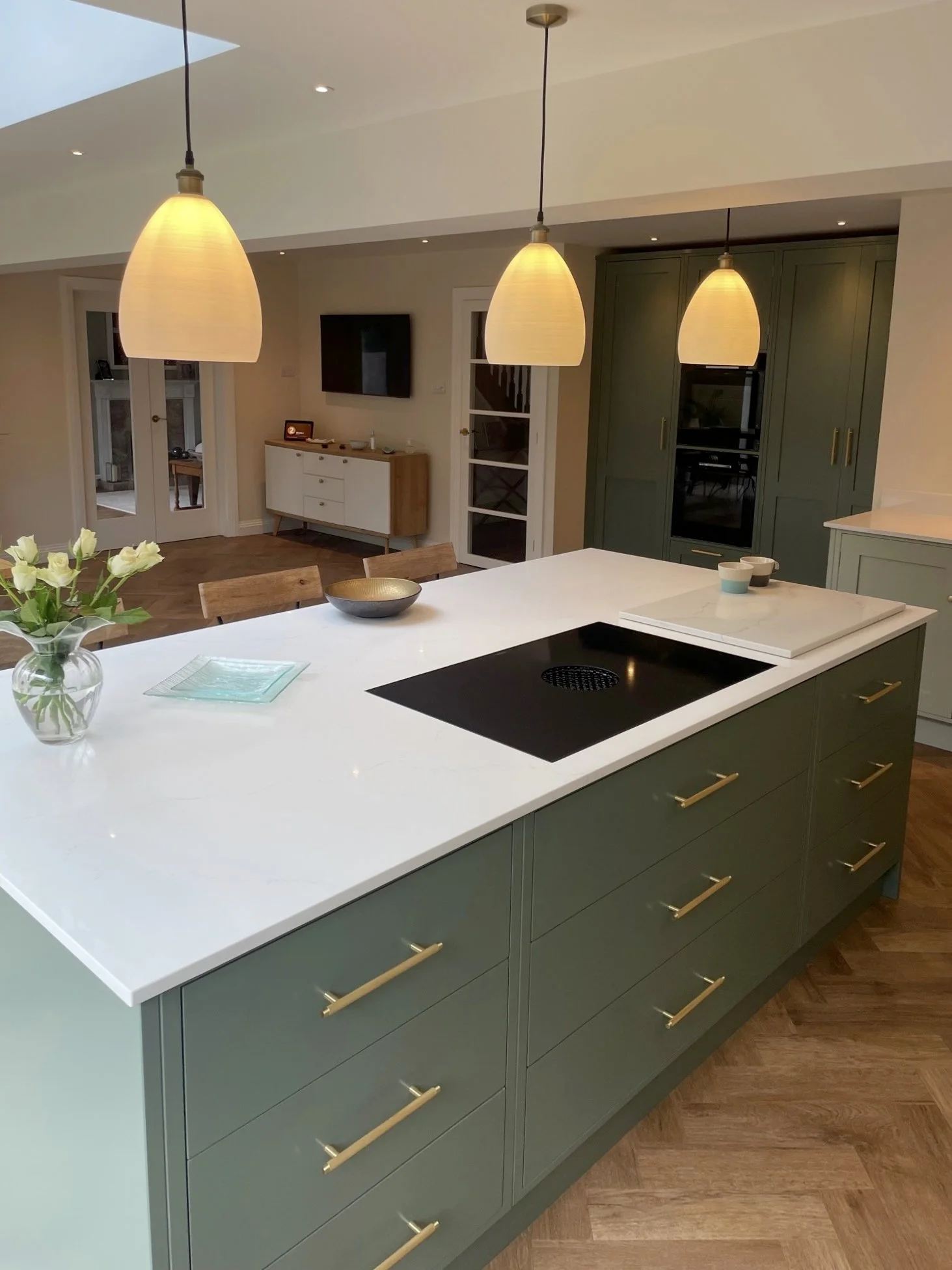 Modern kitchen with green cabinets, white countertop island, hanging pendant lights, and a vase of white flowers.