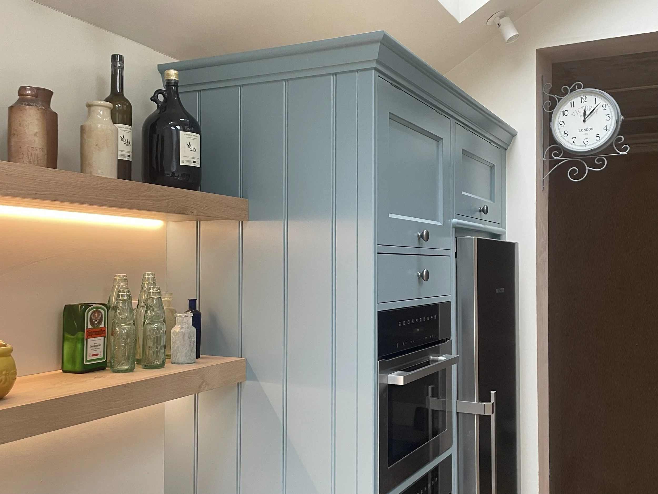 Kitchen corner with blue cabinets, open oven, wall clock, wooden shelves with decorative bottles and jars.