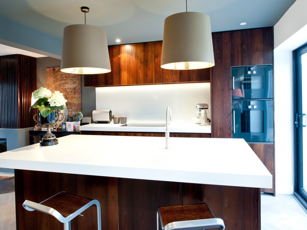 Modern kitchen with white island counter, two large hanging pendant lights, wooden cabinetry, and built-in appliances. There is a vase with white flowers on the counter, and a window/door on the right side.