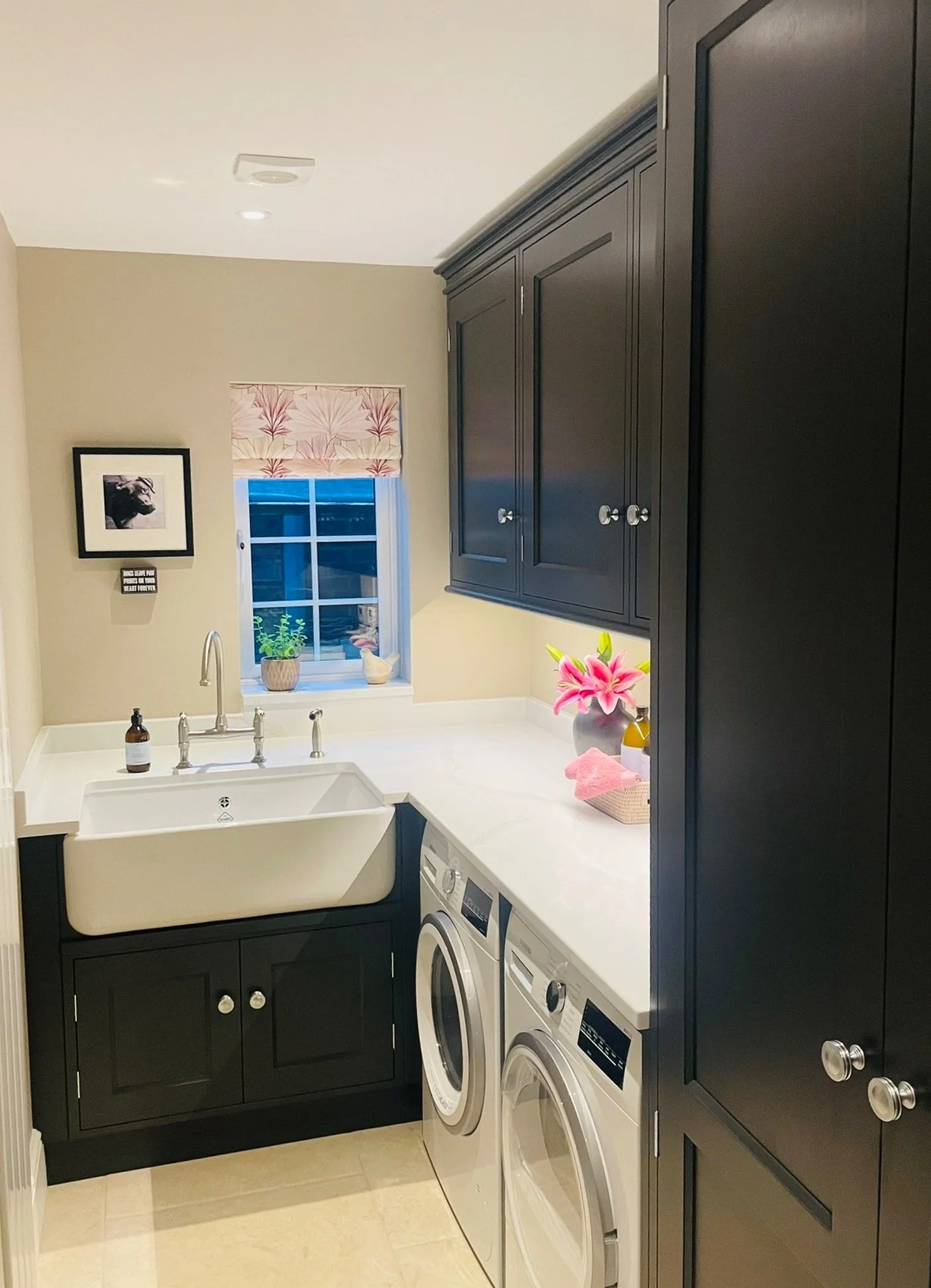 Laundry room with black cabinets, white countertop, a farmhouse sink, and two front-loading washers. There is a window with a pink floral valance, potted plants, pink lilies in a vase, and a framed art piece on the wall.