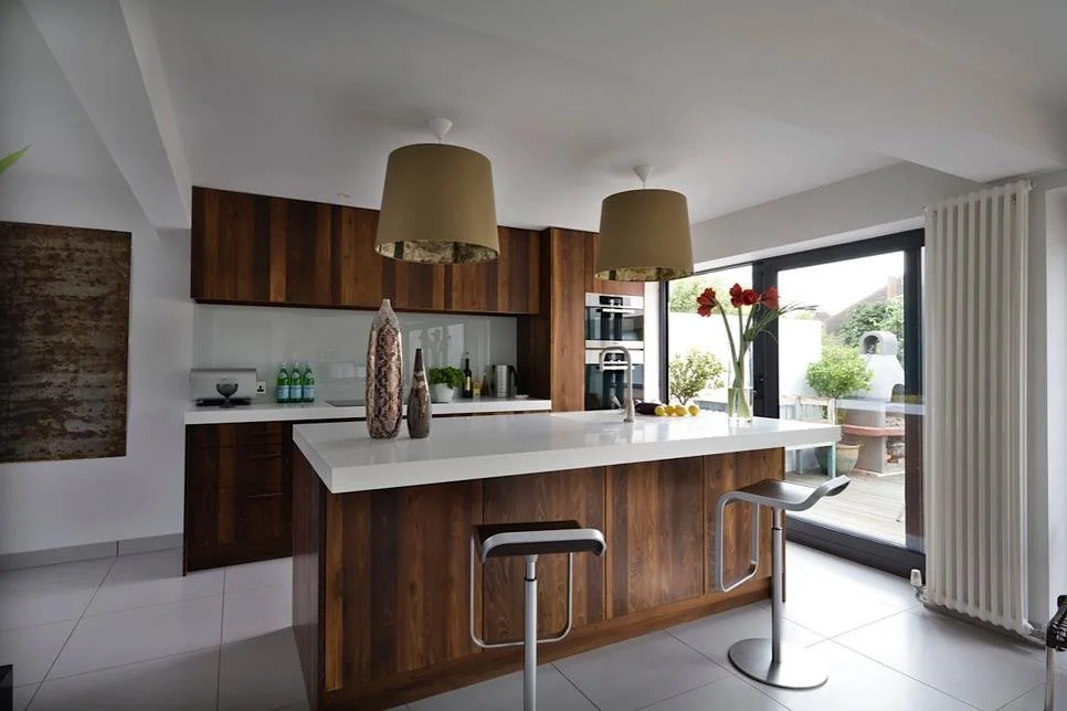 Modern kitchen with wooden cabinets, white countertop island, two beige pendant lights, a large sliding glass door, and a vase with red flowers.