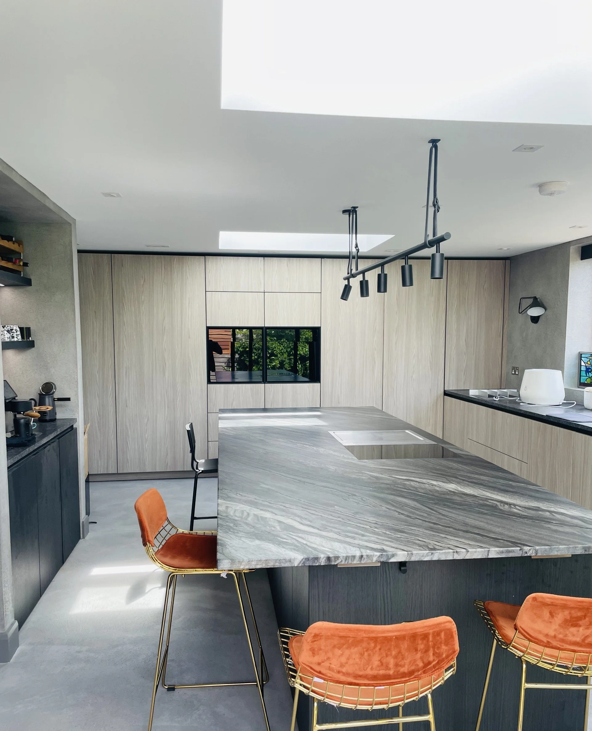 Modern kitchen with a marble island, orange velvet chairs, built-in wooden cabinets, skylights, and minimalist decor.