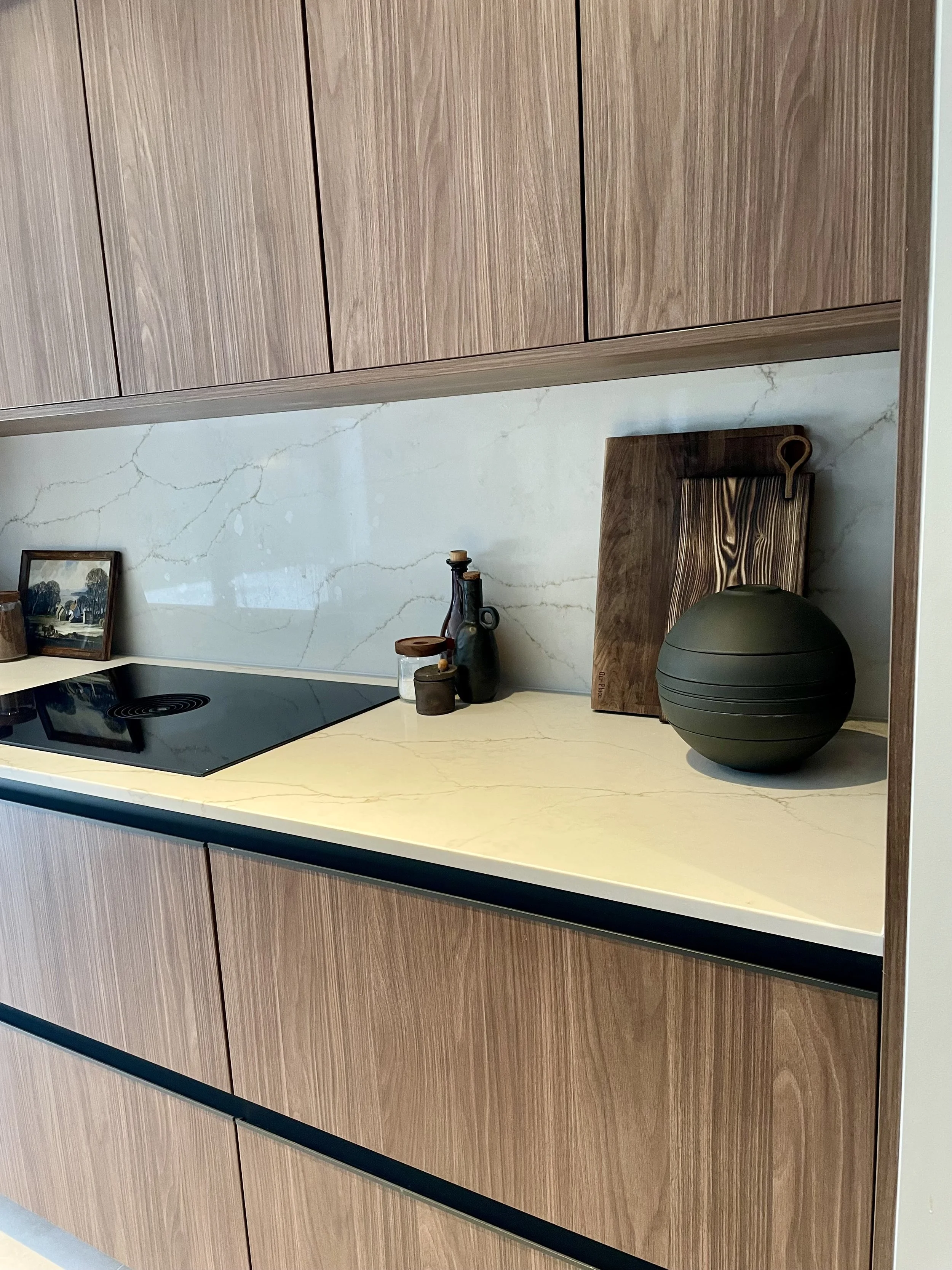 Modern kitchen counter with wooden cabinetry, white marble backsplash, black stove, decorative bottles, a wooden cutting board, a green spherical object, and a framed picture.