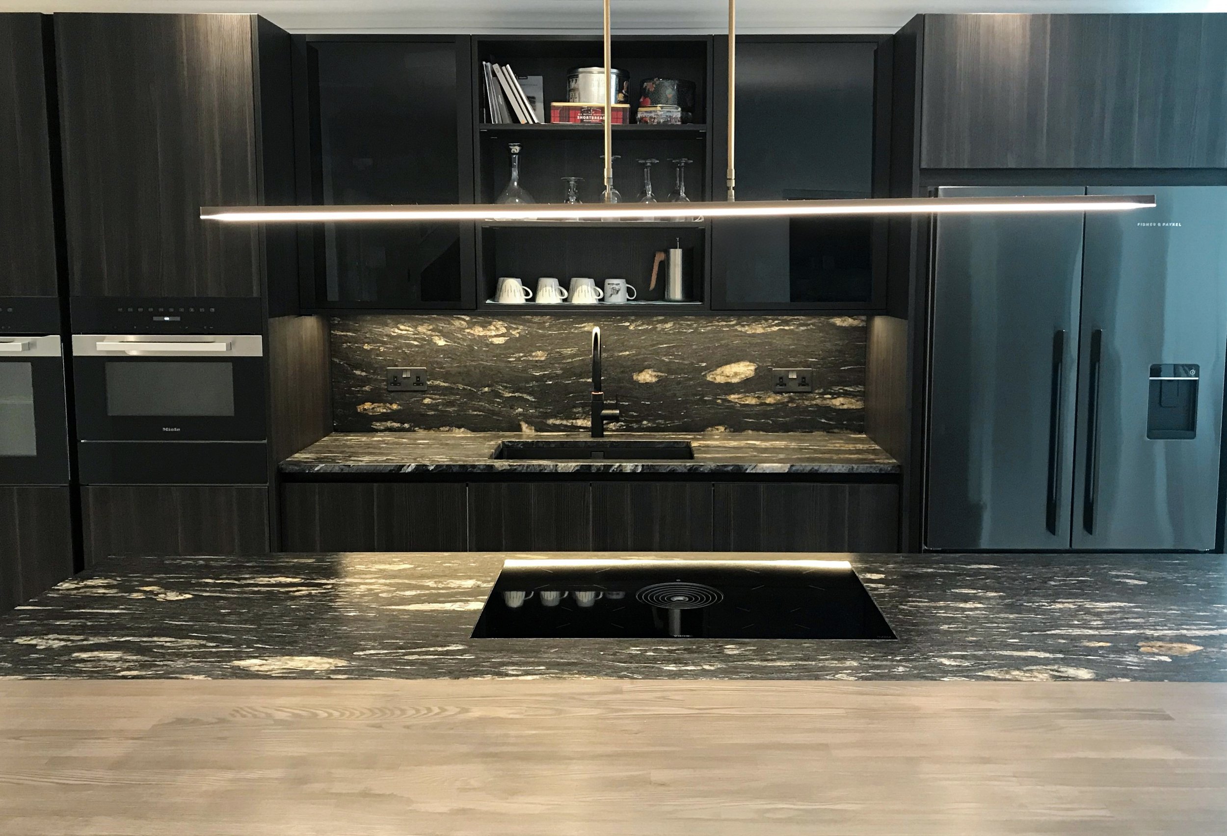Modern kitchen with dark cabinetry, black marble countertops, built-in oven, microwave, and a large refrigerator, with a sleek linear pendant light above the island.
