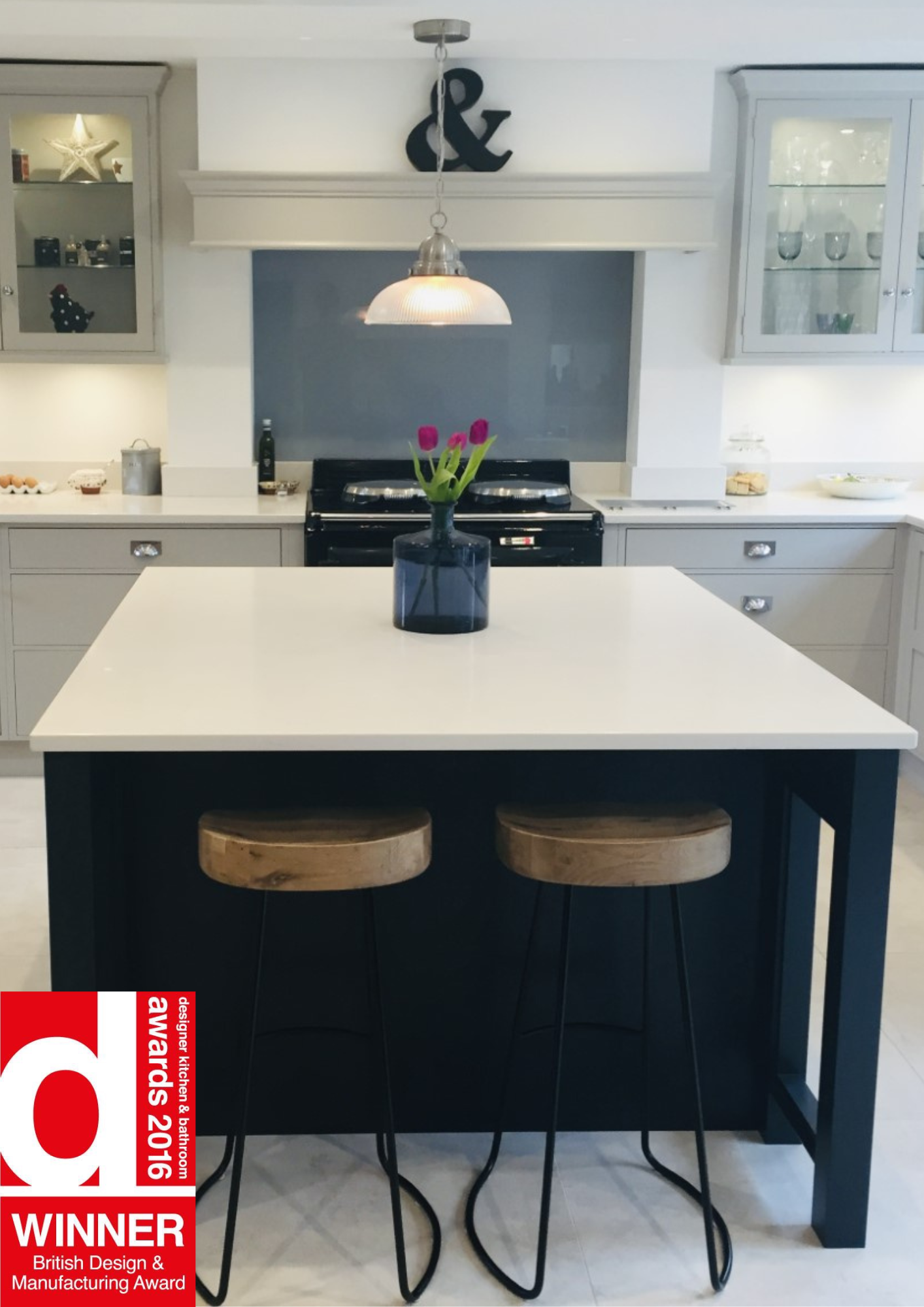 Modern kitchen with a white island, two wooden bar stools, a black vase with pink flowers, and gray cabinetry. An oven is built into the wall, with a decorative ampersand above, and a red award badge is in the lower left corner.
