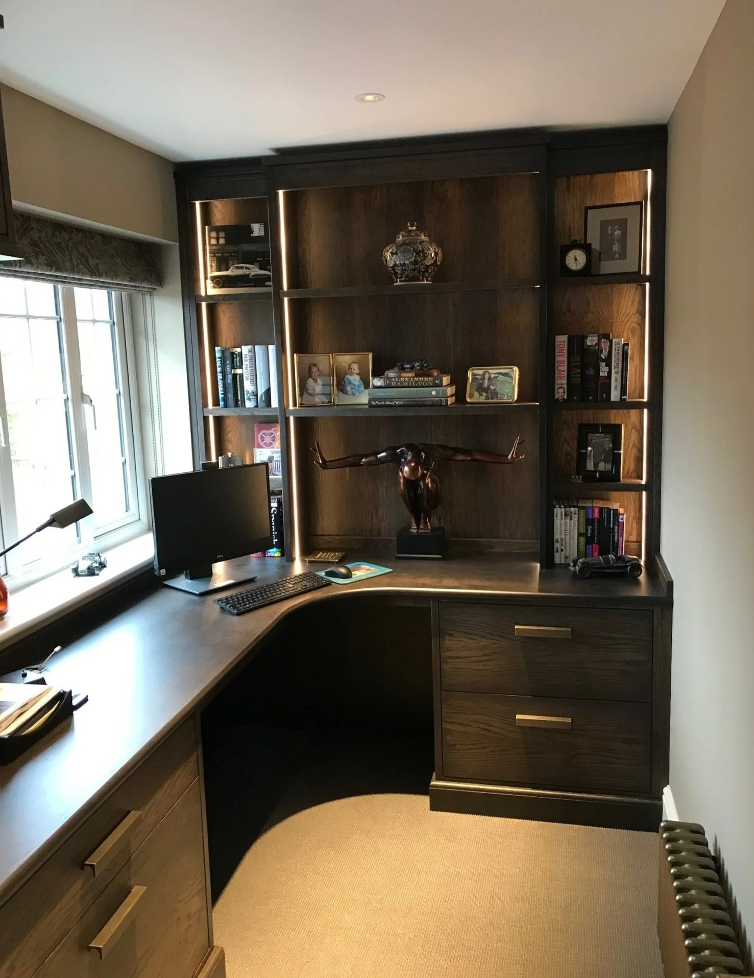 Wooden corner desk with a computer monitor, keyboard, and mouse, set against a window with a valance, built-in shelves with books, photographs, and decorative items, including a bronze sculpture of a man with outstretched arms, and a beige carpeted floor.