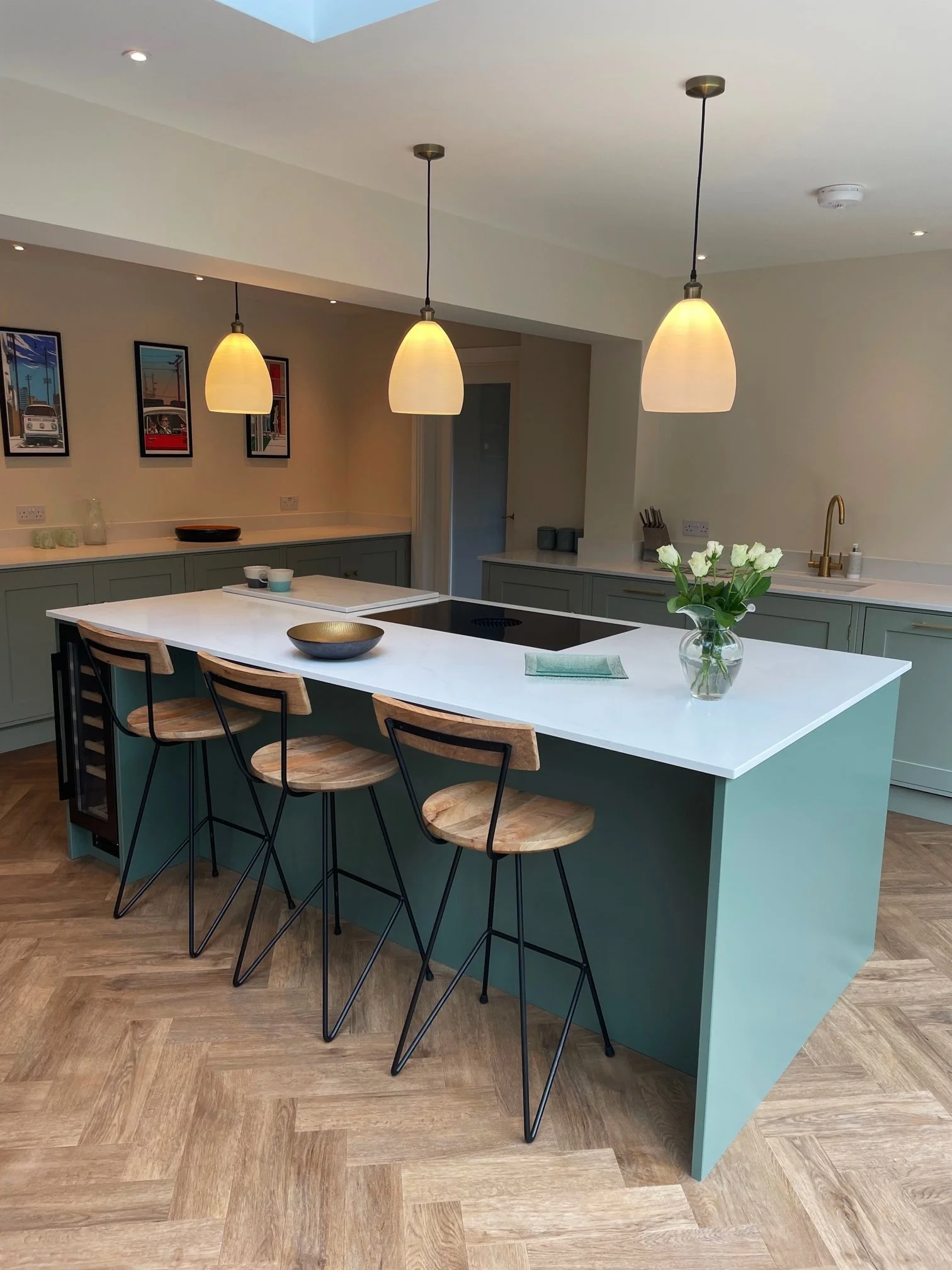 Modern kitchen with a white island countertop, four wooden bar stools, pendant lights, and framed pictures on the wall.