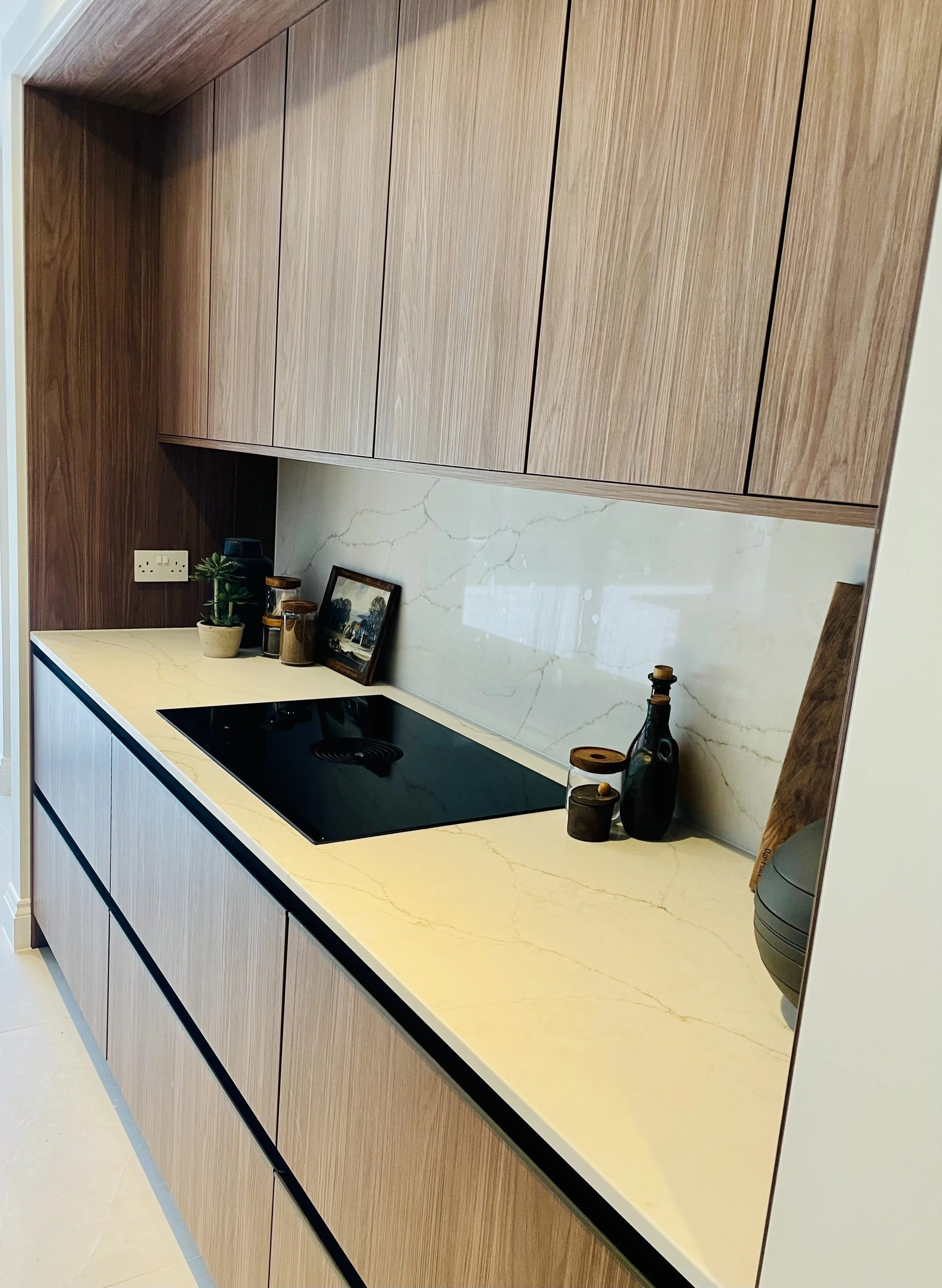Modern kitchen countertop with a black induction stove, decorative bottles, a small frame, and potted plant, with wood cabinets above and a marble backsplash.
