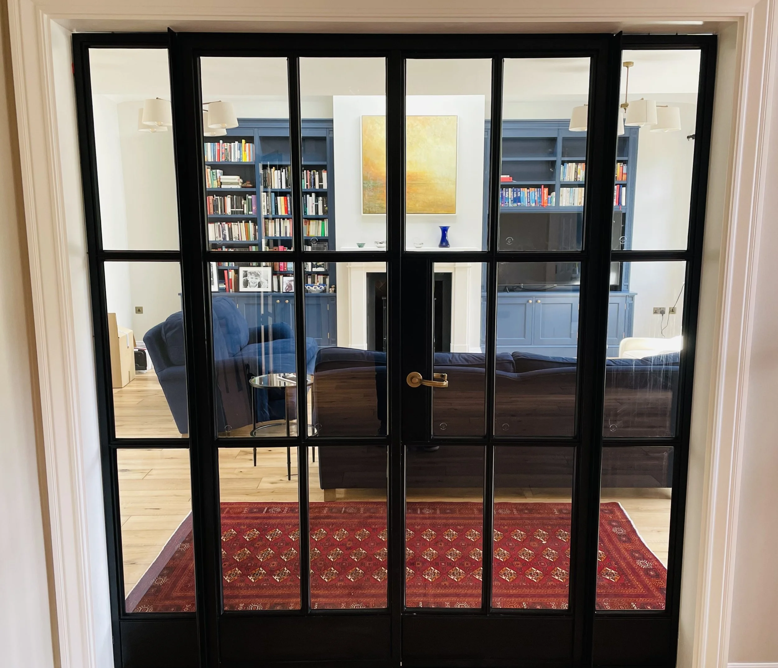 Interior view of a living room seen through black-framed glass doors from an entryway with beige walls and hardwood floors.