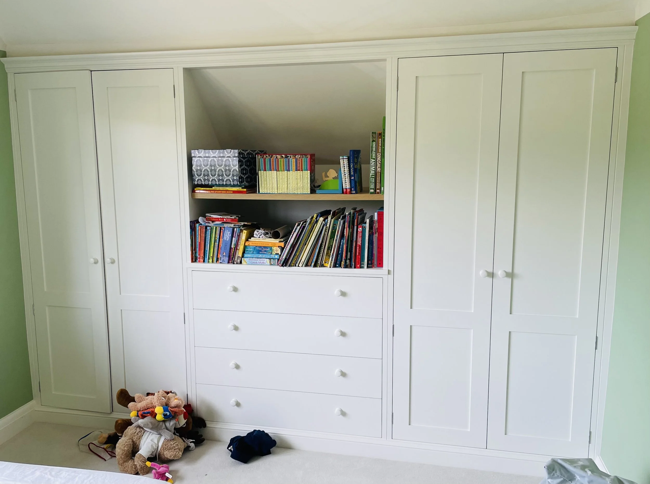 White built-in wardrobe with closed doors and open shelves, filled with books and storage boxes, in a child's bedroom with plush toys on the floor.