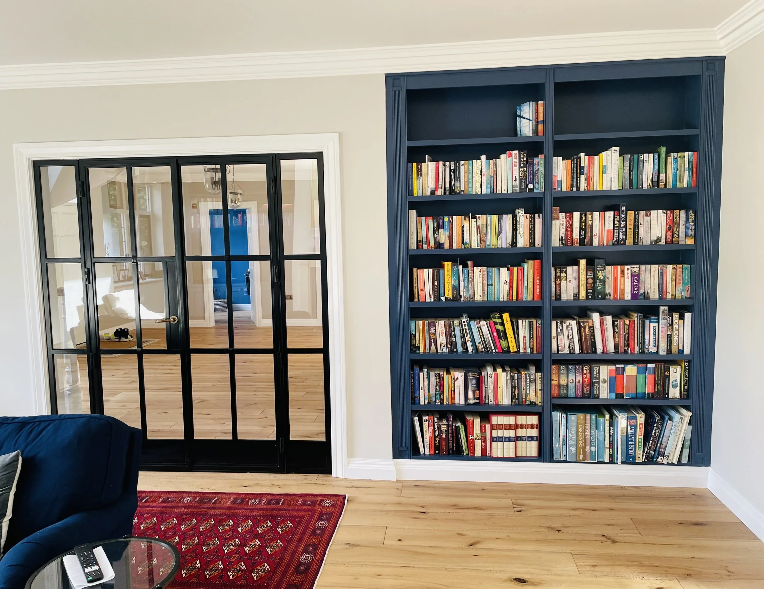 A living room with a large blue bookshelf filled with books, black-framed glass sliding door, hardwood floors, a red patterned rug, dark blue sofa, and a glass side table with remote controls.