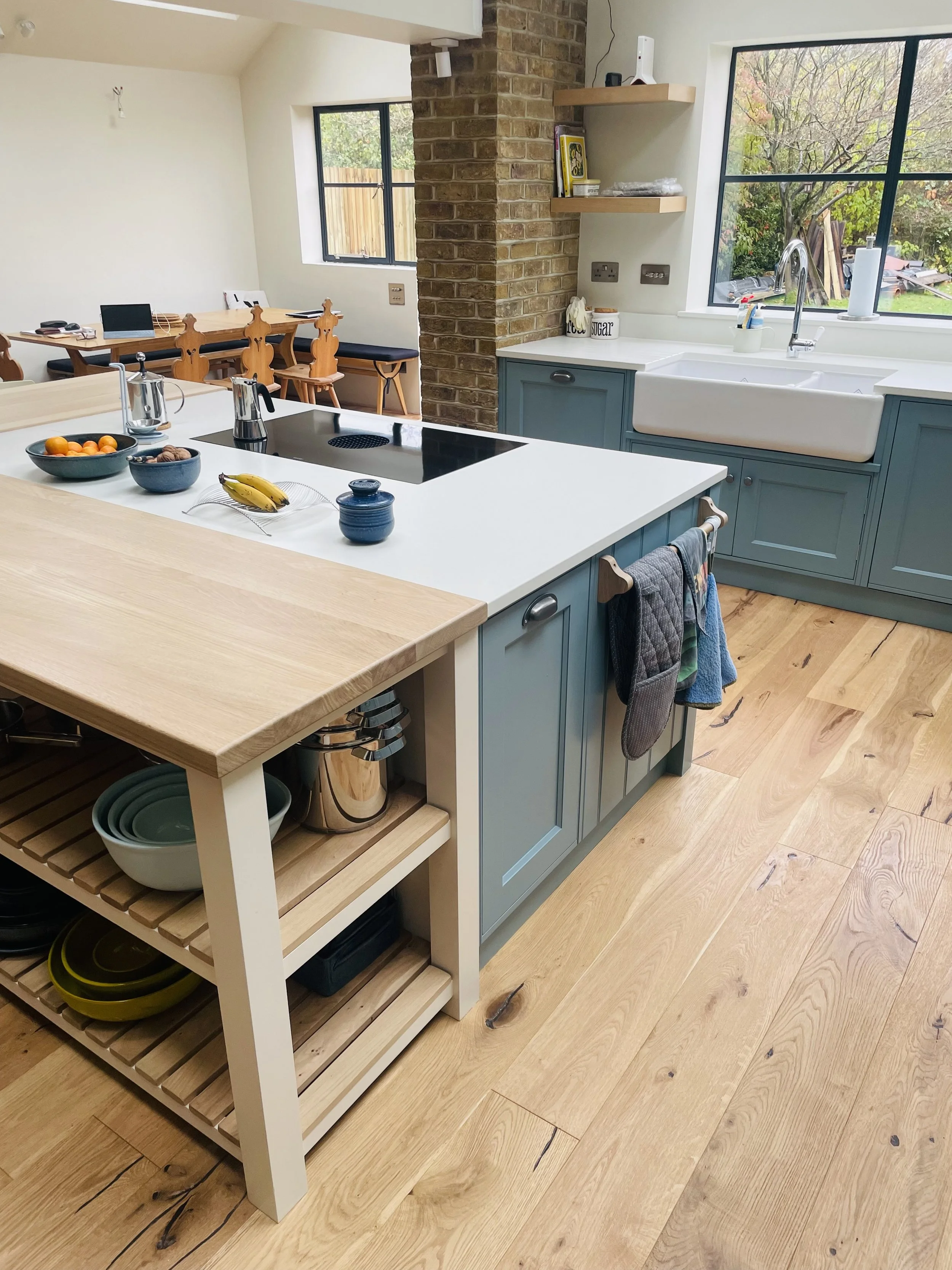 Modern kitchen with a central island featuring a white countertop, blue cabinets, and open shelves. There is a sink under a large window, and a brick column dividing the kitchen and dining area. The dining area has a wooden table and chairs. The floo