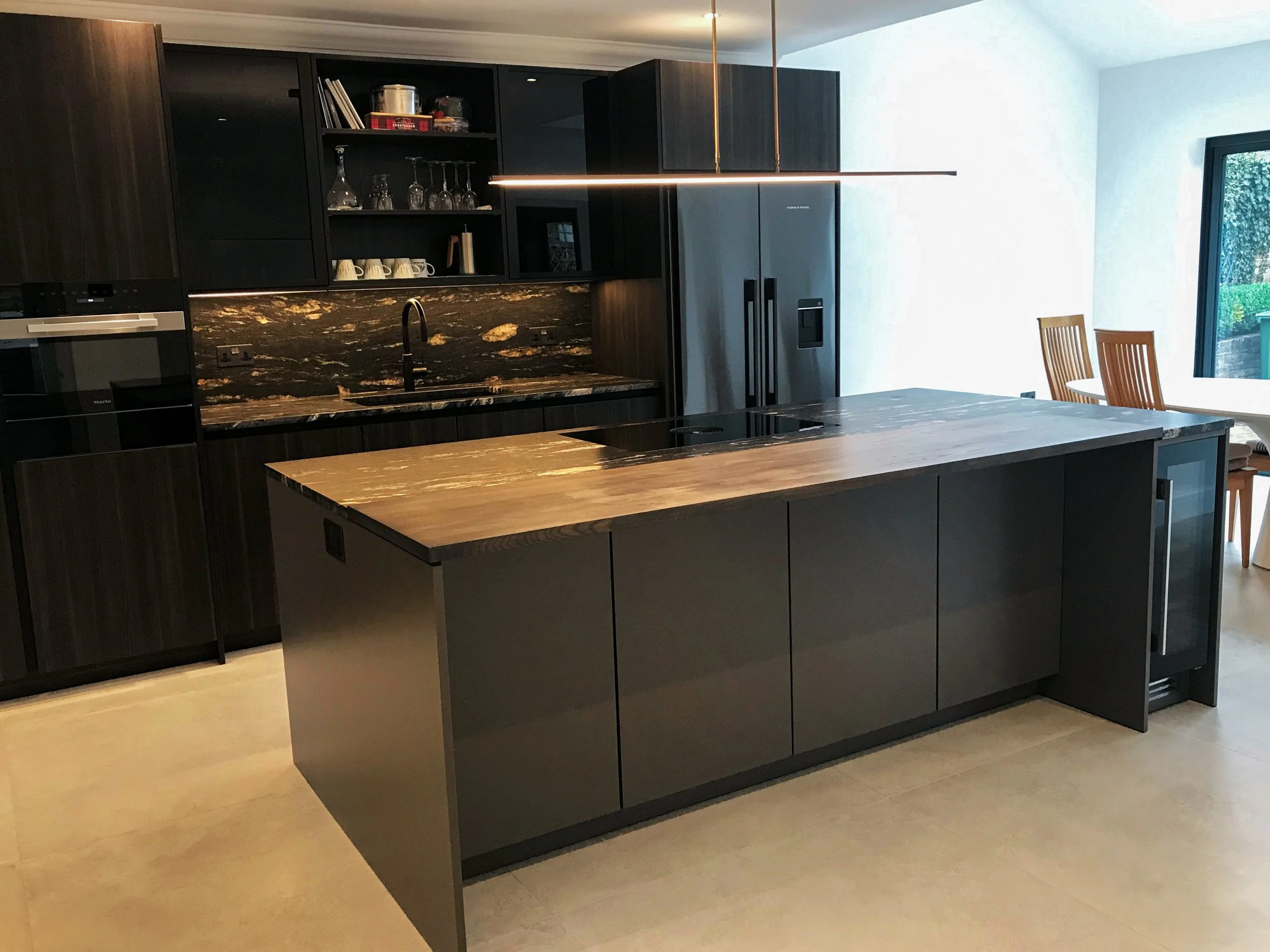 Modern kitchen with black cabinetry, a wooden kitchen island, a black marble backsplash, and a stainless steel refrigerator. There is a window with a view outside and a dining table with chairs in the background.