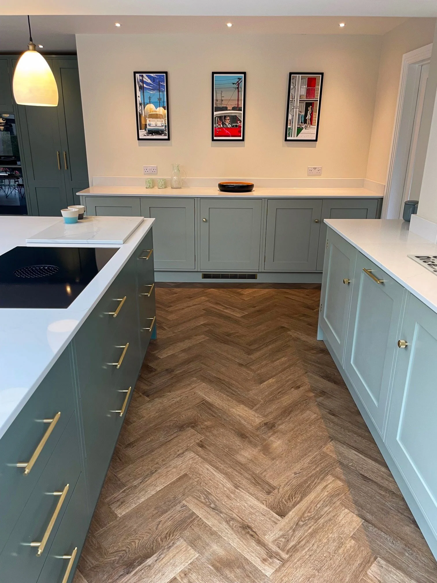 A modern kitchen with green lower cabinets, white countertops, and a wooden herringbone floor. There are three framed art prints on the wall and a pendant light hanging from the ceiling.