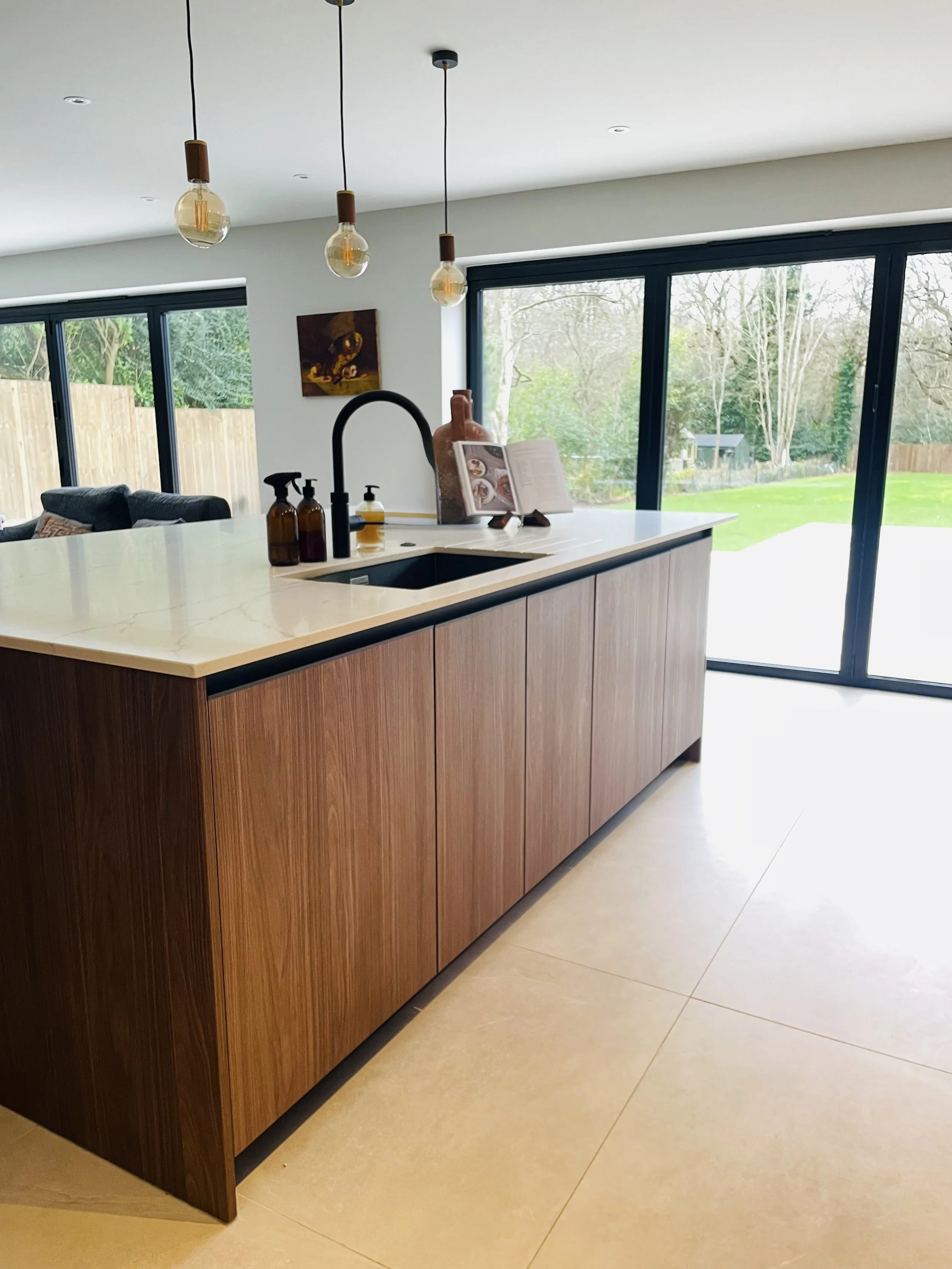 Modern kitchen island with a marble top and wooden cabinets, black faucet, soap dispensers, decorative vase, and open cookbook on a stand, with large sliding glass doors leading to a backyard.