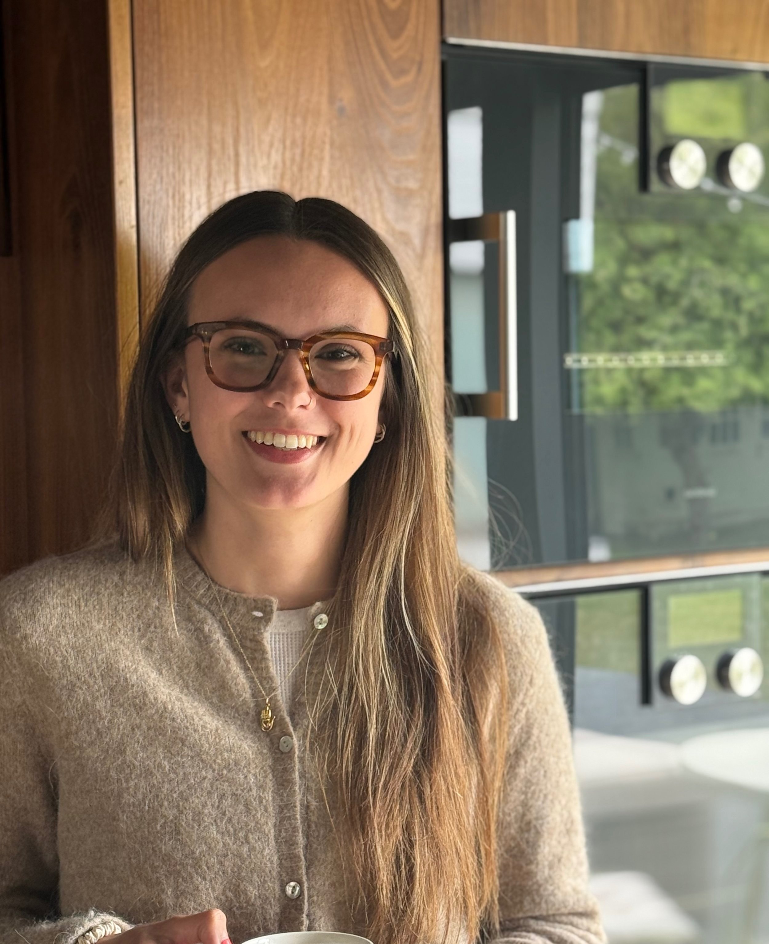 A woman with long brown hair, wearing glasses and a beige cardigan, smiling indoors near a wooden wall and kitchen appliances.