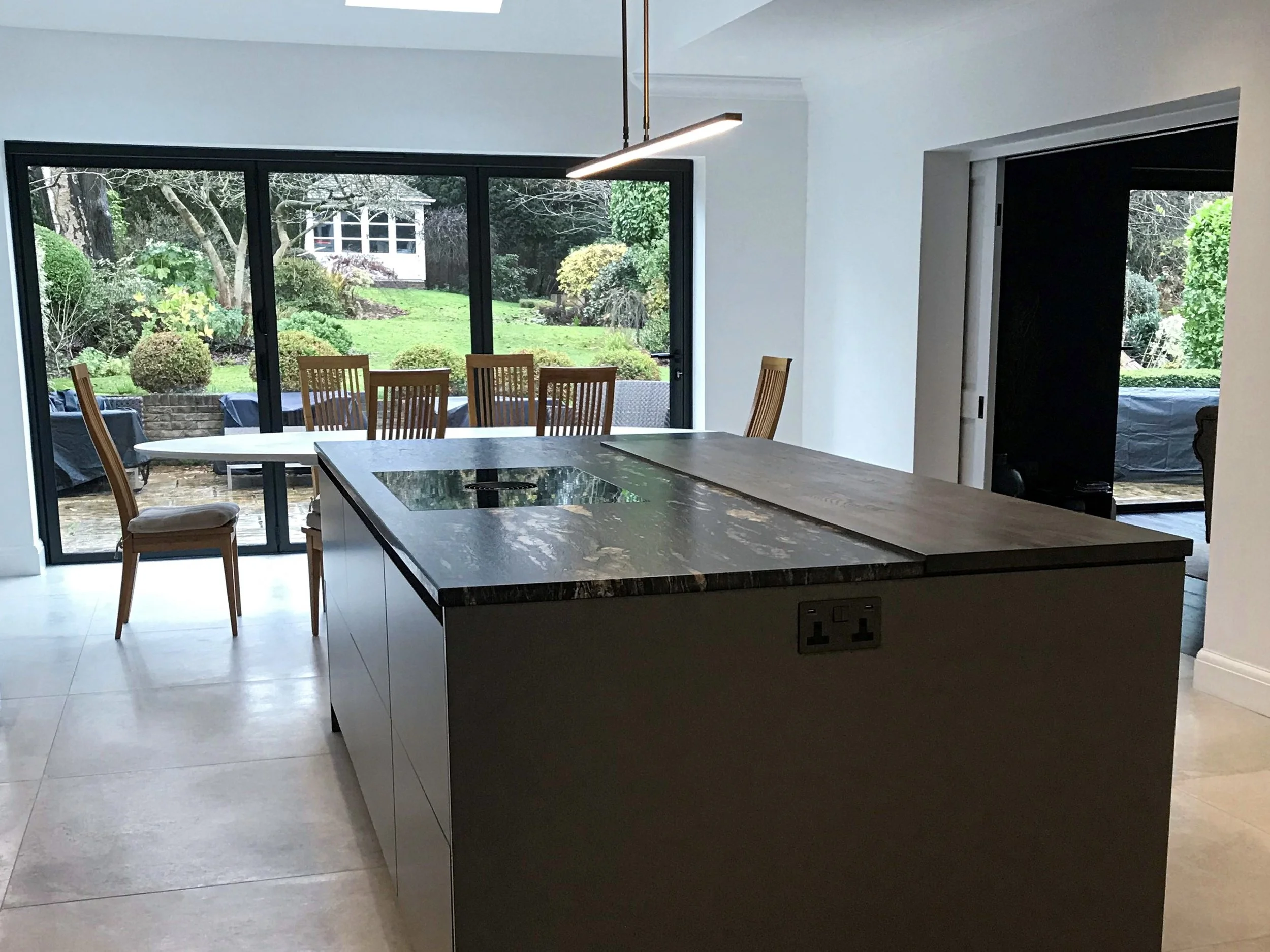 Modern kitchen with dark marble island, dining area with wooden chairs, large sliding glass doors opening to a lush backyard with trees and shrubs.