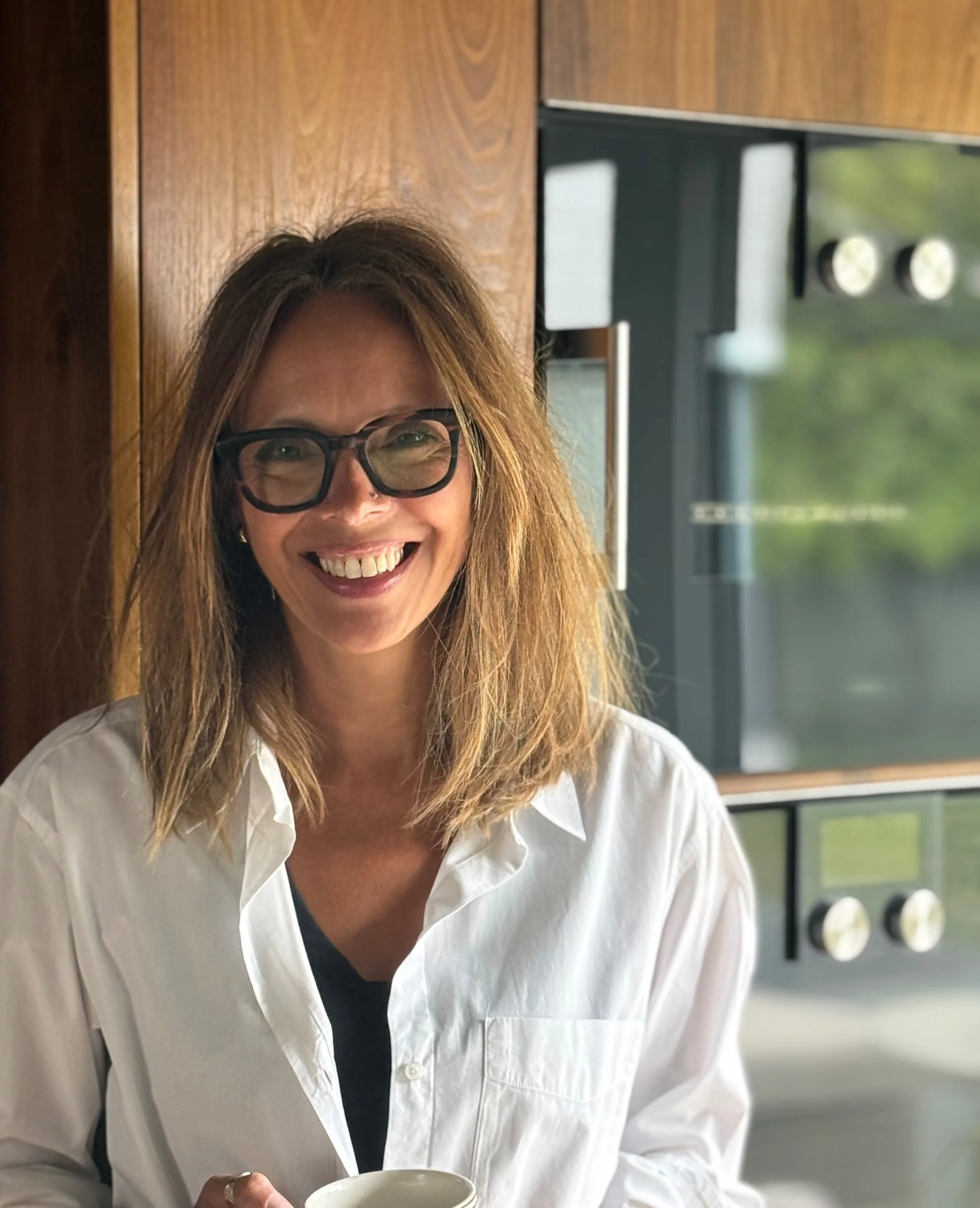 A woman with shoulder-length brown hair, glasses, and a white button-up shirt smiling while holding a coffee mug in a modern kitchen.