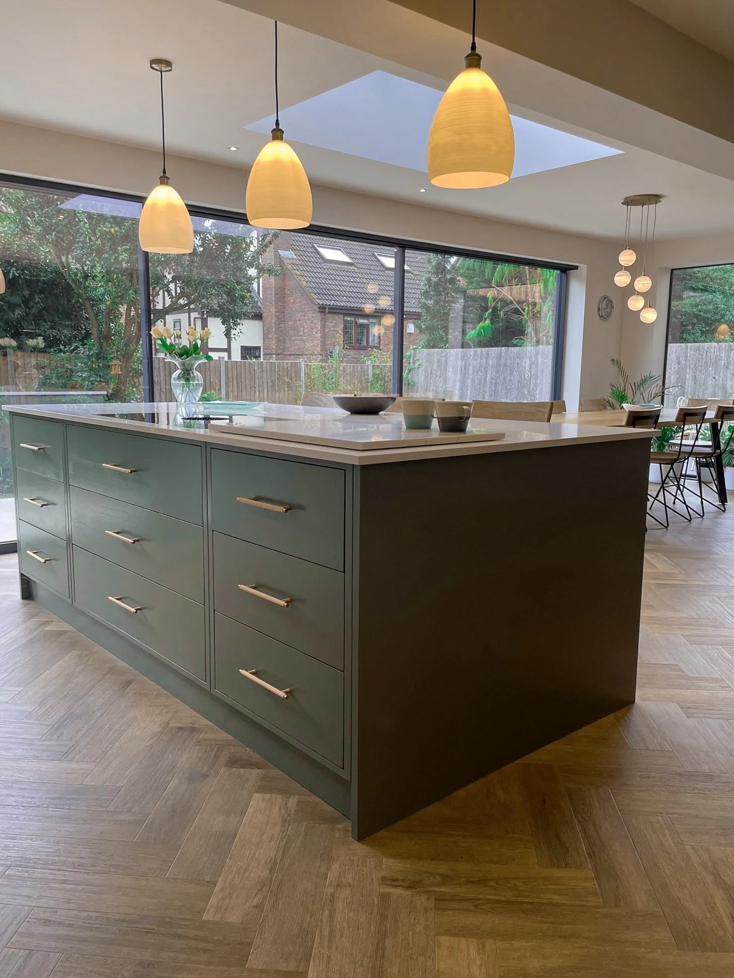 Modern kitchen with green island, pendant lights, large windows, and wooden flooring.