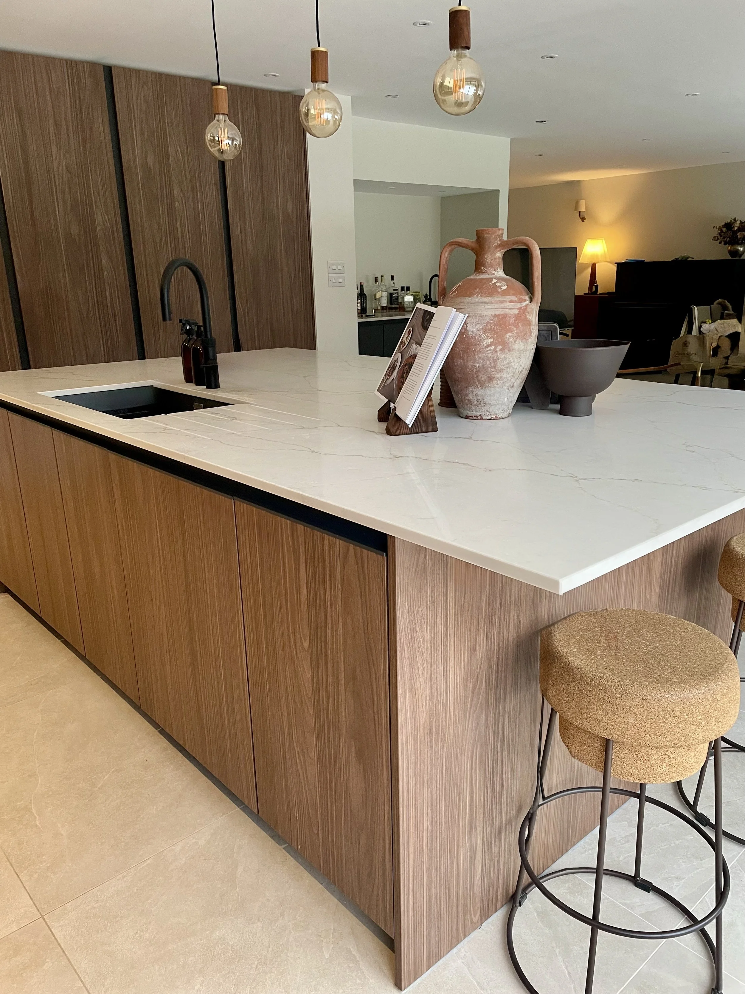 Modern kitchen island with a white marble countertop, wooden cabinetry, and decorative items including a large terracotta vase, a black bowl, and a cookbook stand. Three hanging pendant lights are above the island, and a bar stool with a cushioned se