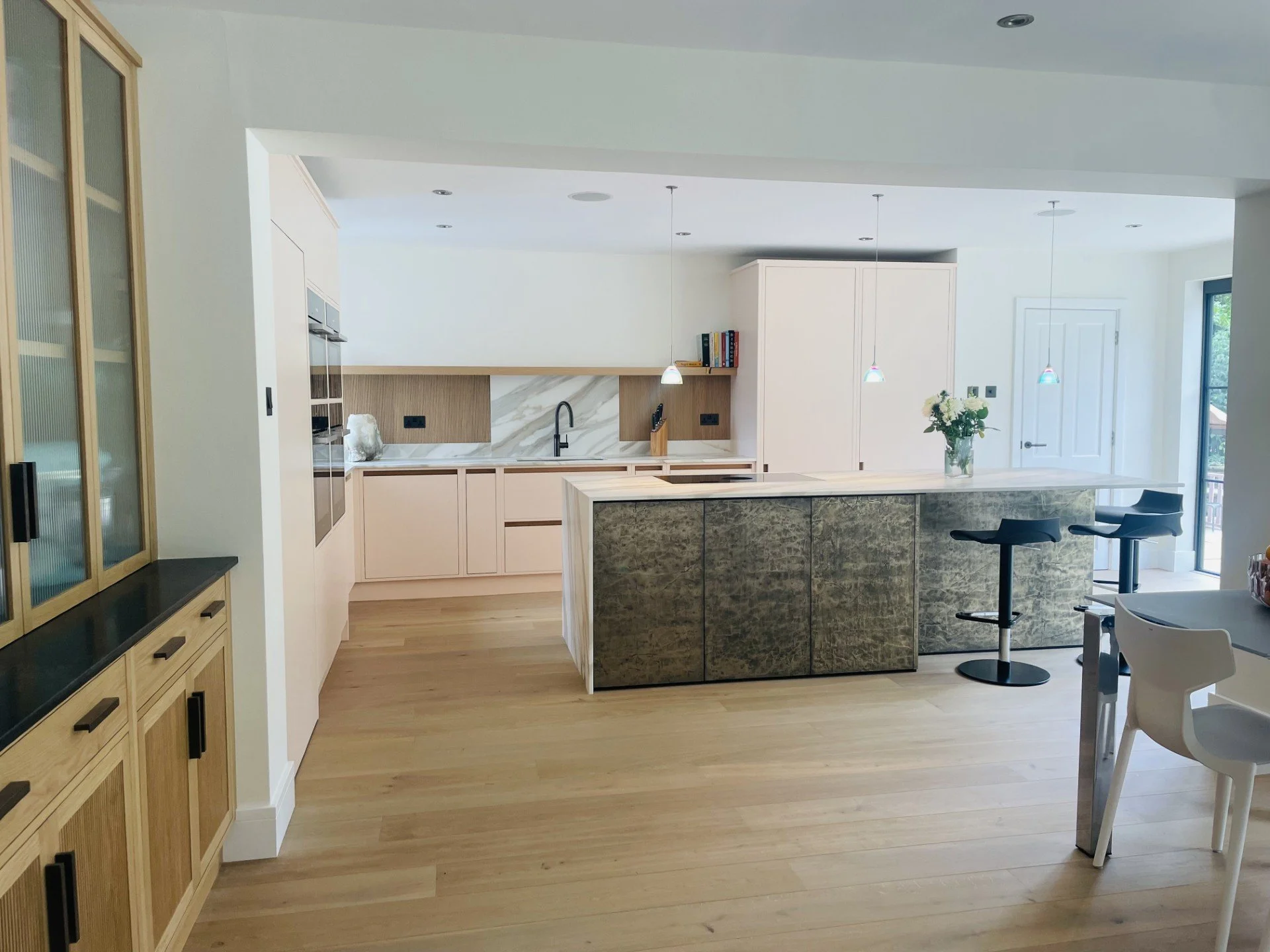 Modern kitchen with light wood cabinets, a marble backsplash, an island with a green stone front, and a vase with white flowers on the countertop. There are hanging pendant lights and black bar stools at the island.