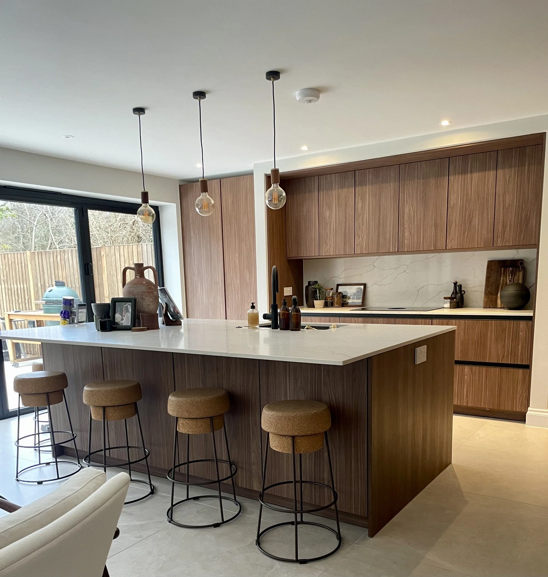 Modern kitchen with wooden cabinets, white marble backsplash, and a kitchen island with a white countertop and four beige barstools. There are hanging pendant lights above the island, a sliding glass door leading outside, and decorative items on the counter.
