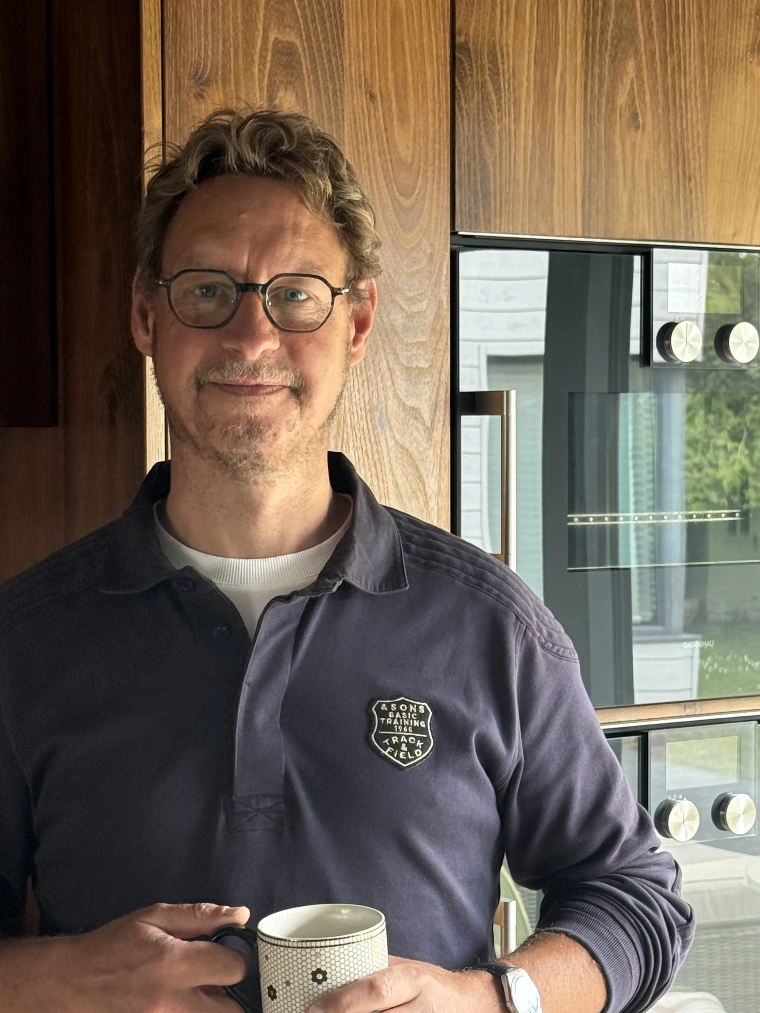 A man with glasses and a beard holding a mug, standing in front of wooden kitchen cabinets and a modern oven.
