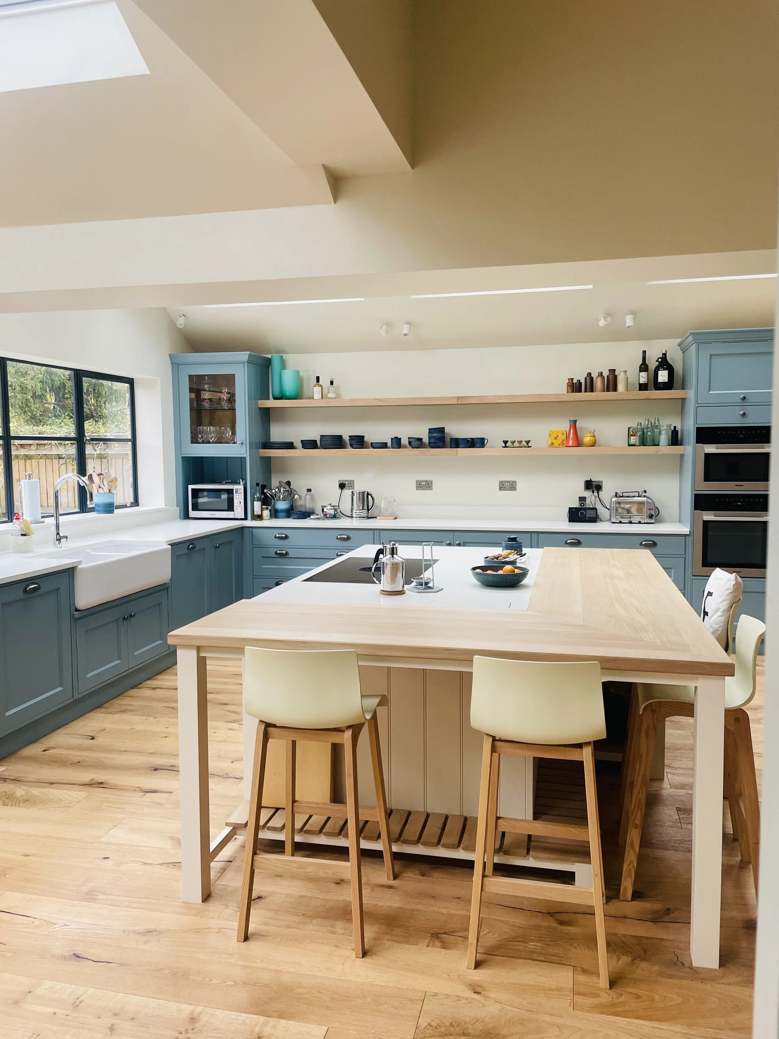 Modern kitchen with blue cabinetry, a central island with a wooden top, and a high sloped ceiling with natural light, featuring open shelves with dishes and bottles, and various small appliances.
