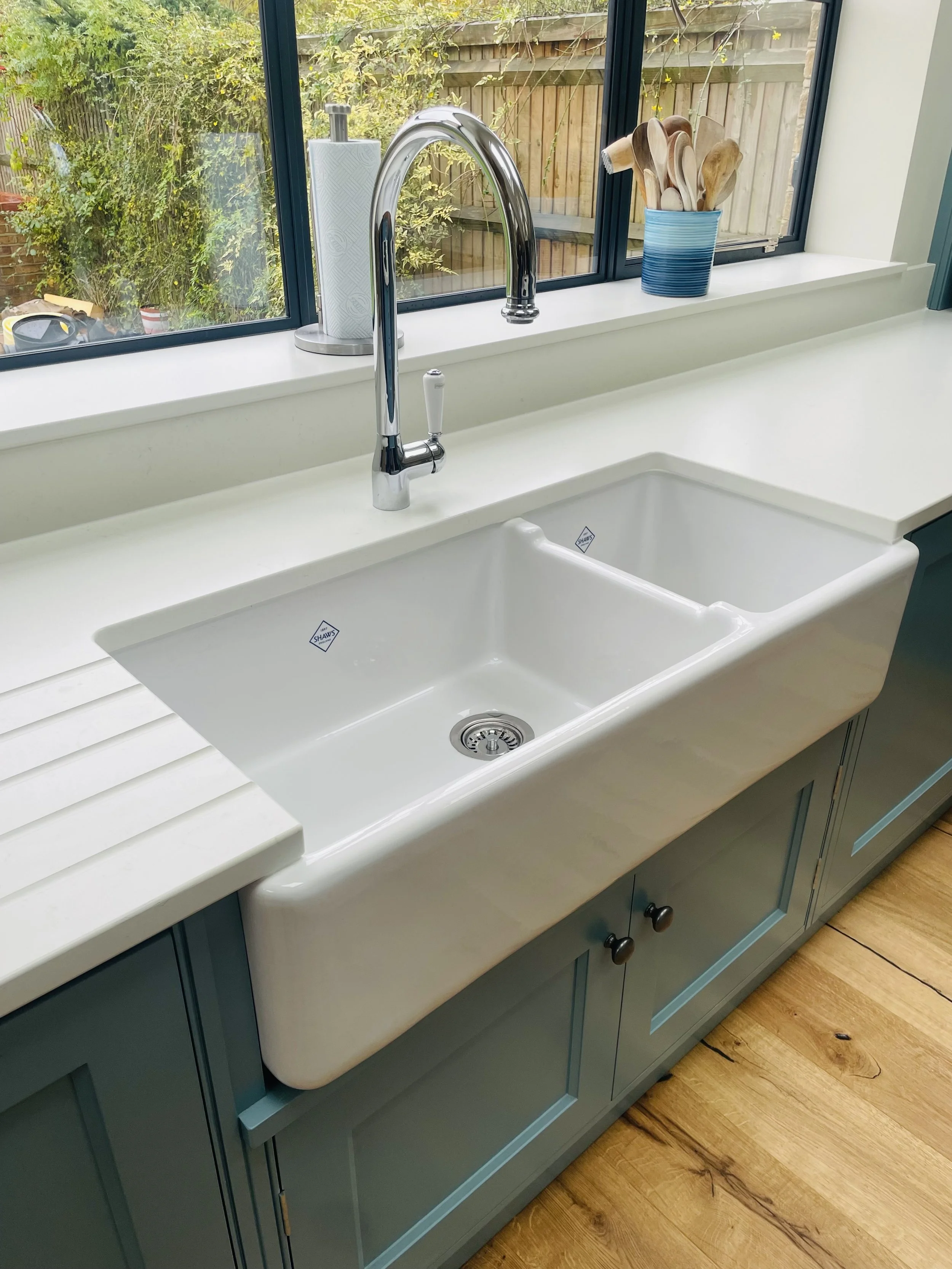 Kitchen sink with a white farmhouse style basin, chrome faucet, placed on a white countertop next to a window, with a paper towel holder and a container of wooden utensils on the windowsill.