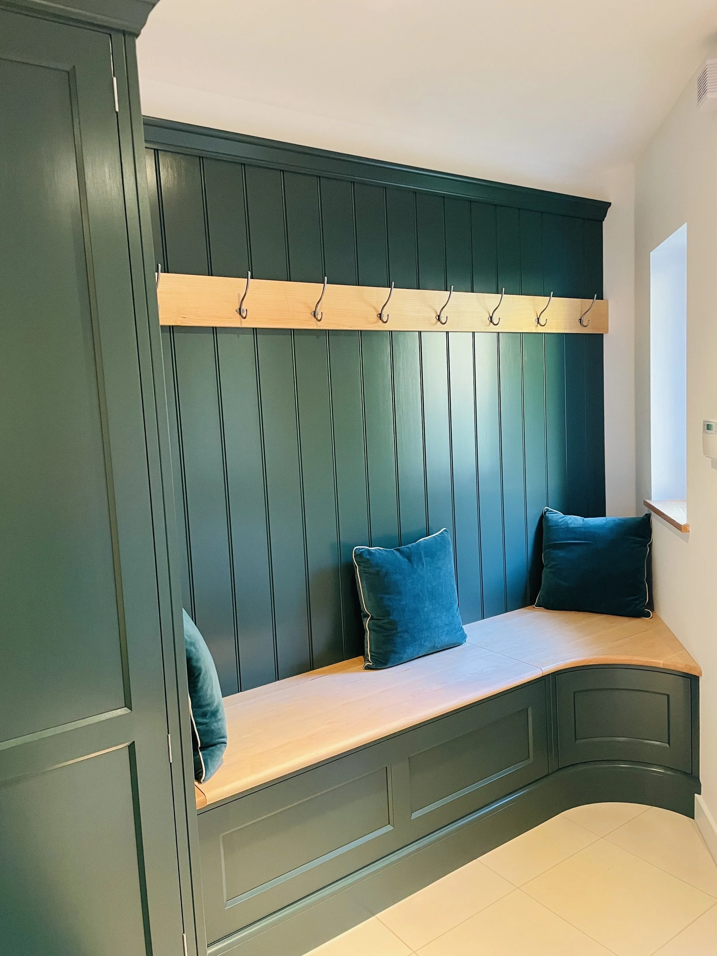 Green built-in corner bench with a wooden seat, teal pillows, a wooden coat rack with metal hooks above, and a small window on a white wall.