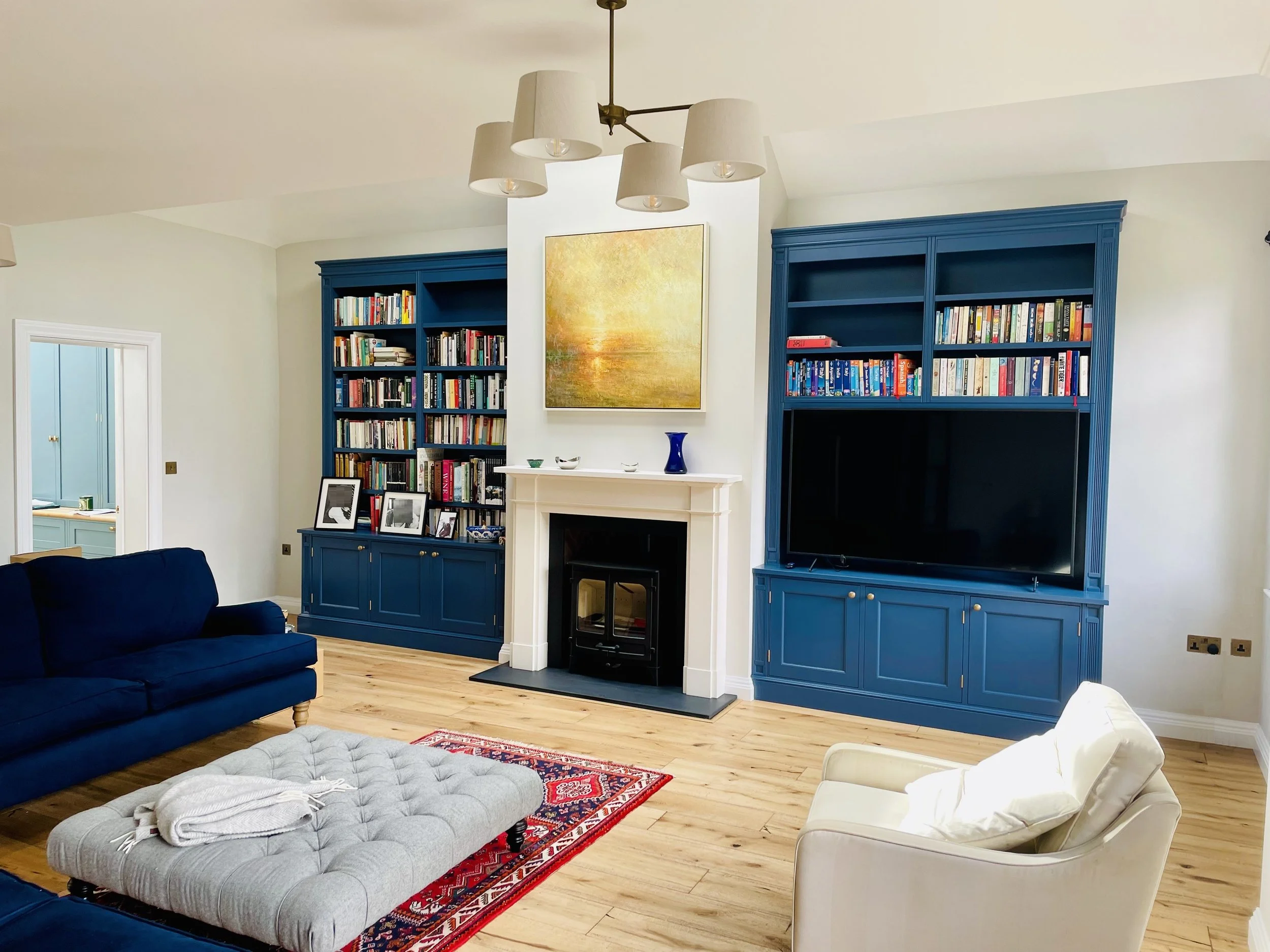 Living room with blue built-in bookshelves, a white fireplace, a beige armchair, a dark blue sofa, a light-colored ottoman, a patterned rug, and a recent abstract painting above the fireplace.