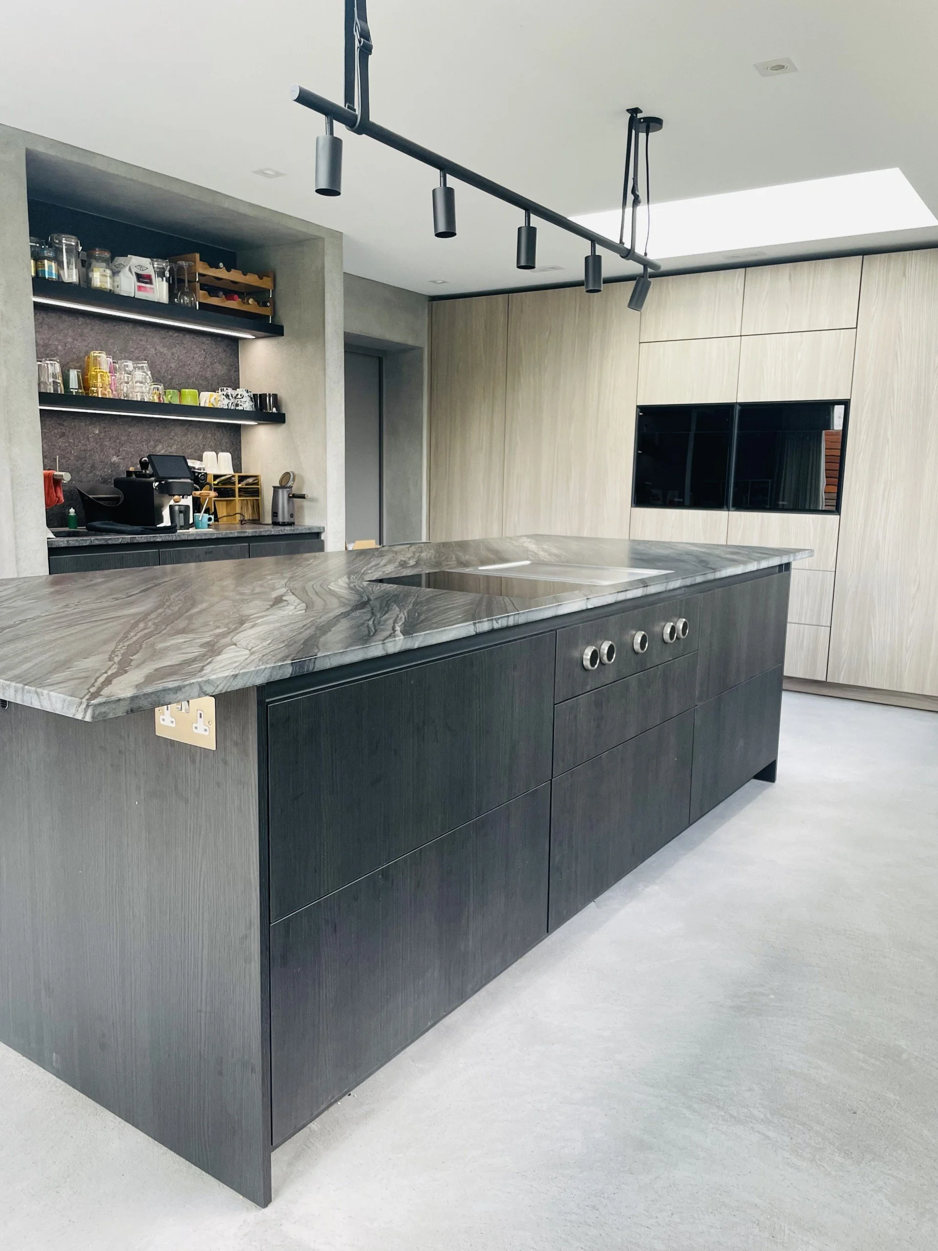Modern kitchen with black and gray cabinetry, marble countertop, open shelving with glassware, built-in black appliances, and track lighting.