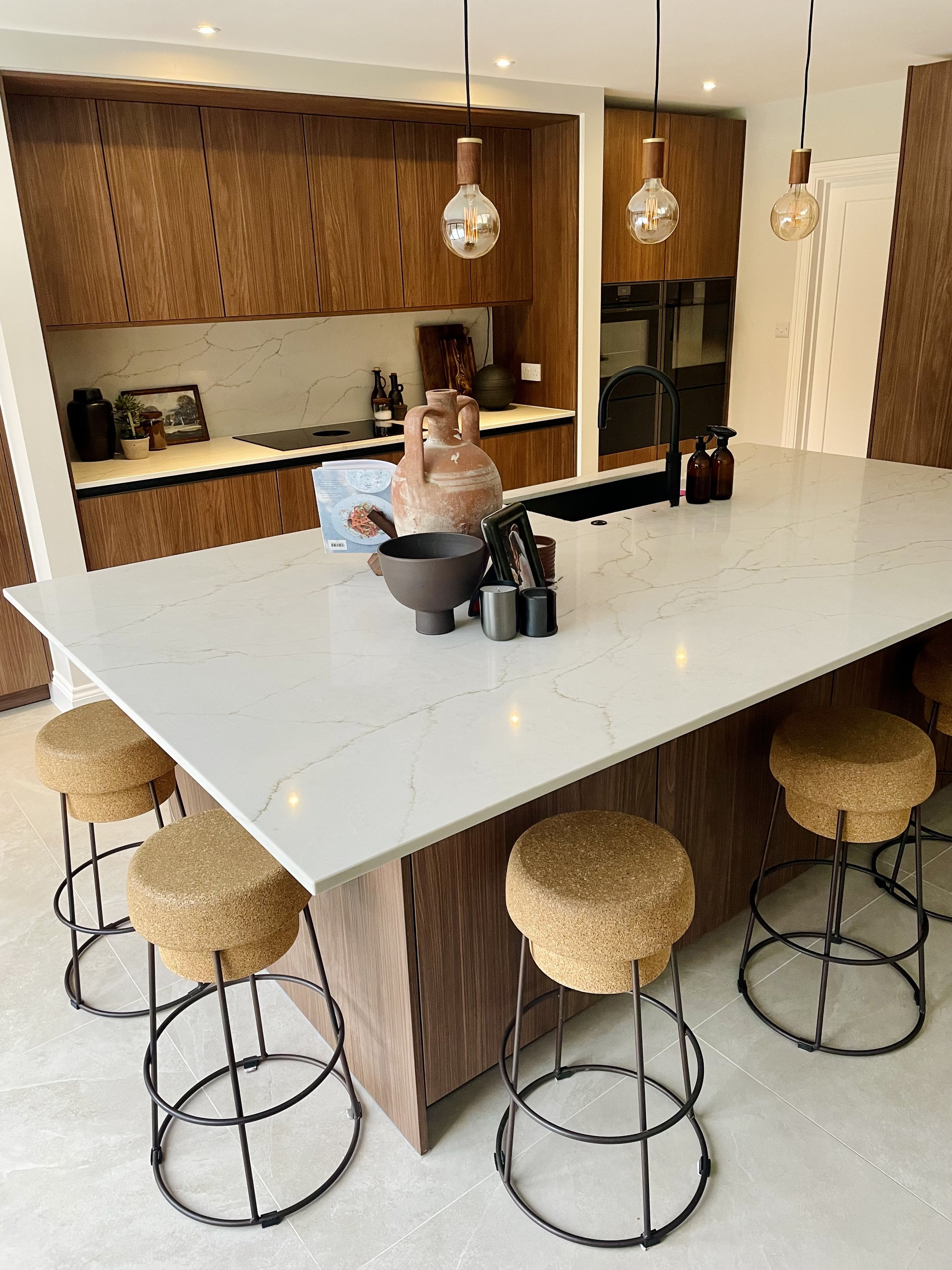 Modern kitchen with a white marble island, four beige cushioned bar stools with black metal legs, wooden cabinetry, pendant lights, and black appliances.