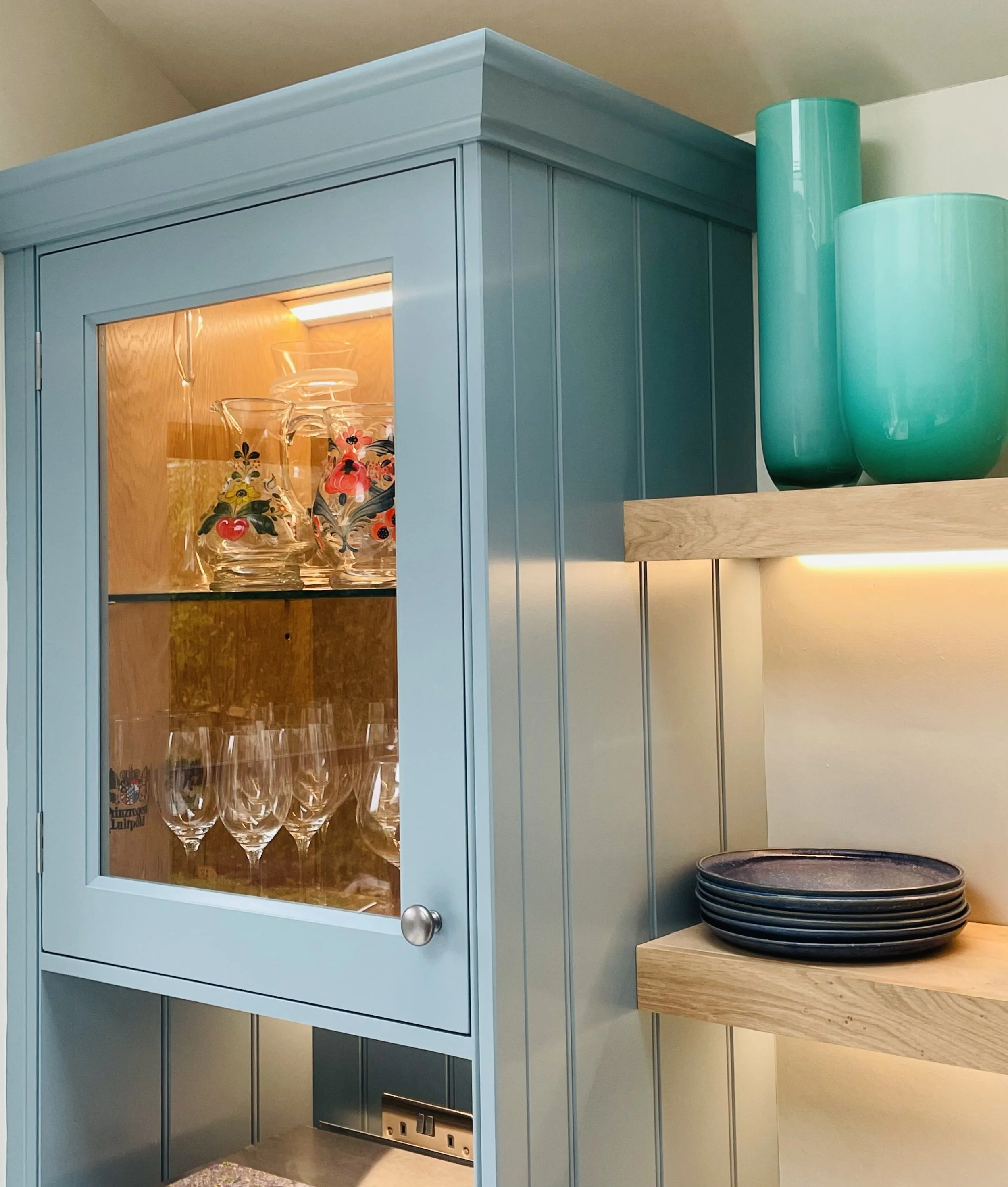 A light blue cabinet with glass doors displaying glassware and colorful painted floral designs inside. To the right, two teal vases and a stack of black plates are on wooden shelves.