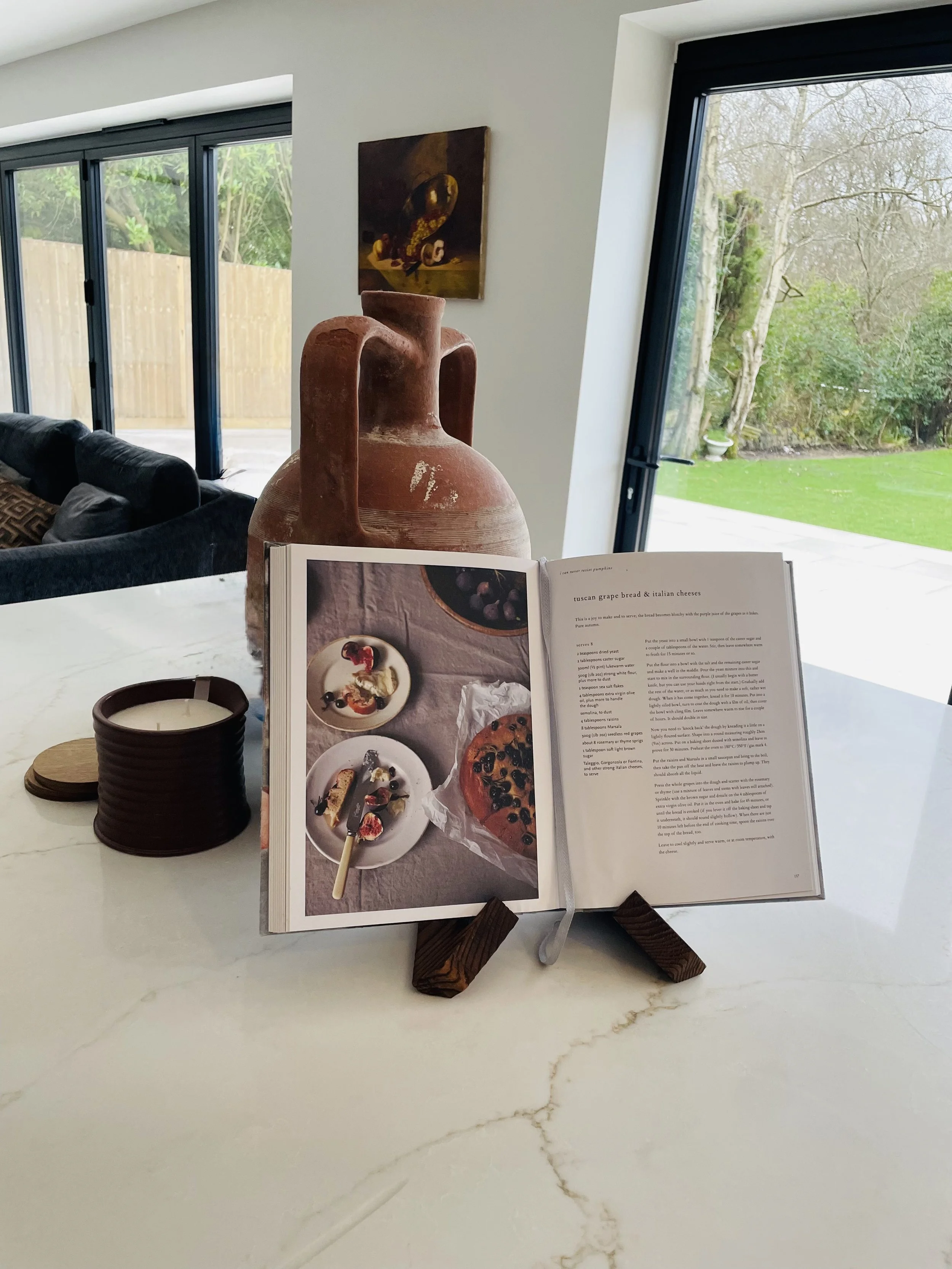 Open cookbook on a stand, displaying recipes and food photos, on a white marble kitchen counter. Behind it is a large terracotta vase, with a black sofa and sliding glass doors revealing a backyard with trees and grass.