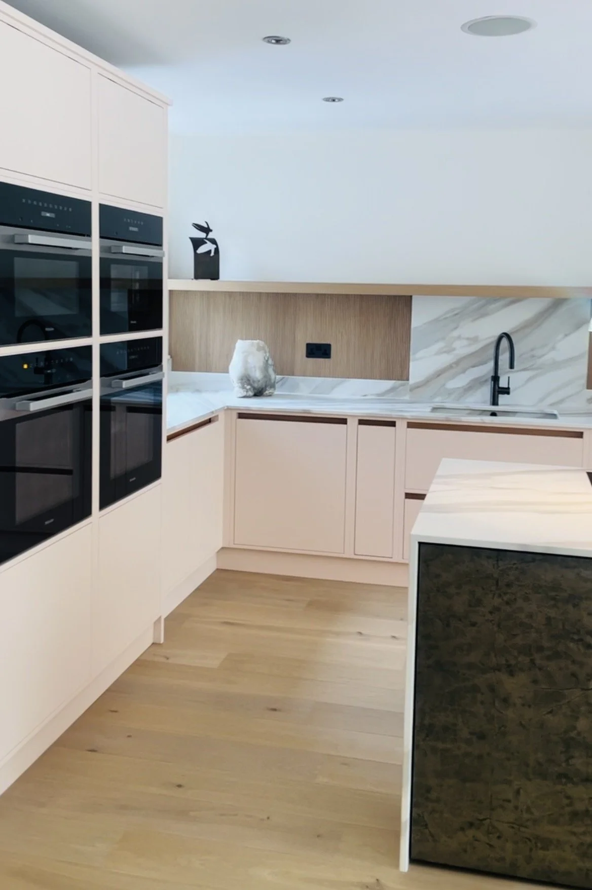 Modern kitchen with beige cabinets, black faucet, marble backsplash, built-in black appliances, light wooden flooring, and decorative items on the counter.