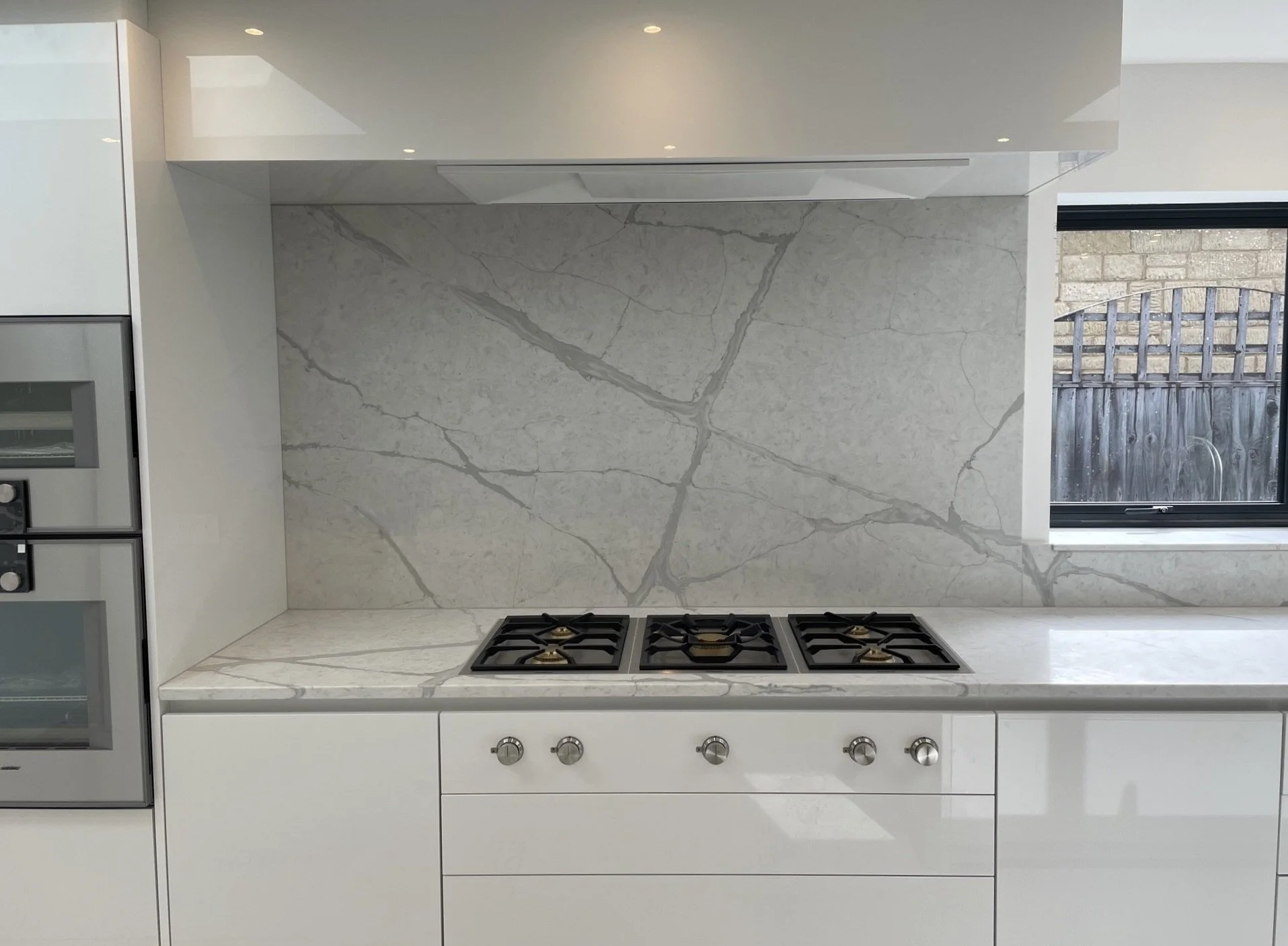 Modern kitchen with a white marble backsplash and countertop, built-in oven, and a black window with a wooden fence outside.