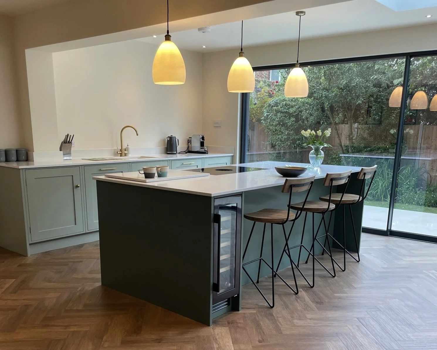 Modern kitchen with a green island, three bar stools, light fixture, and a large window showing a garden outside.