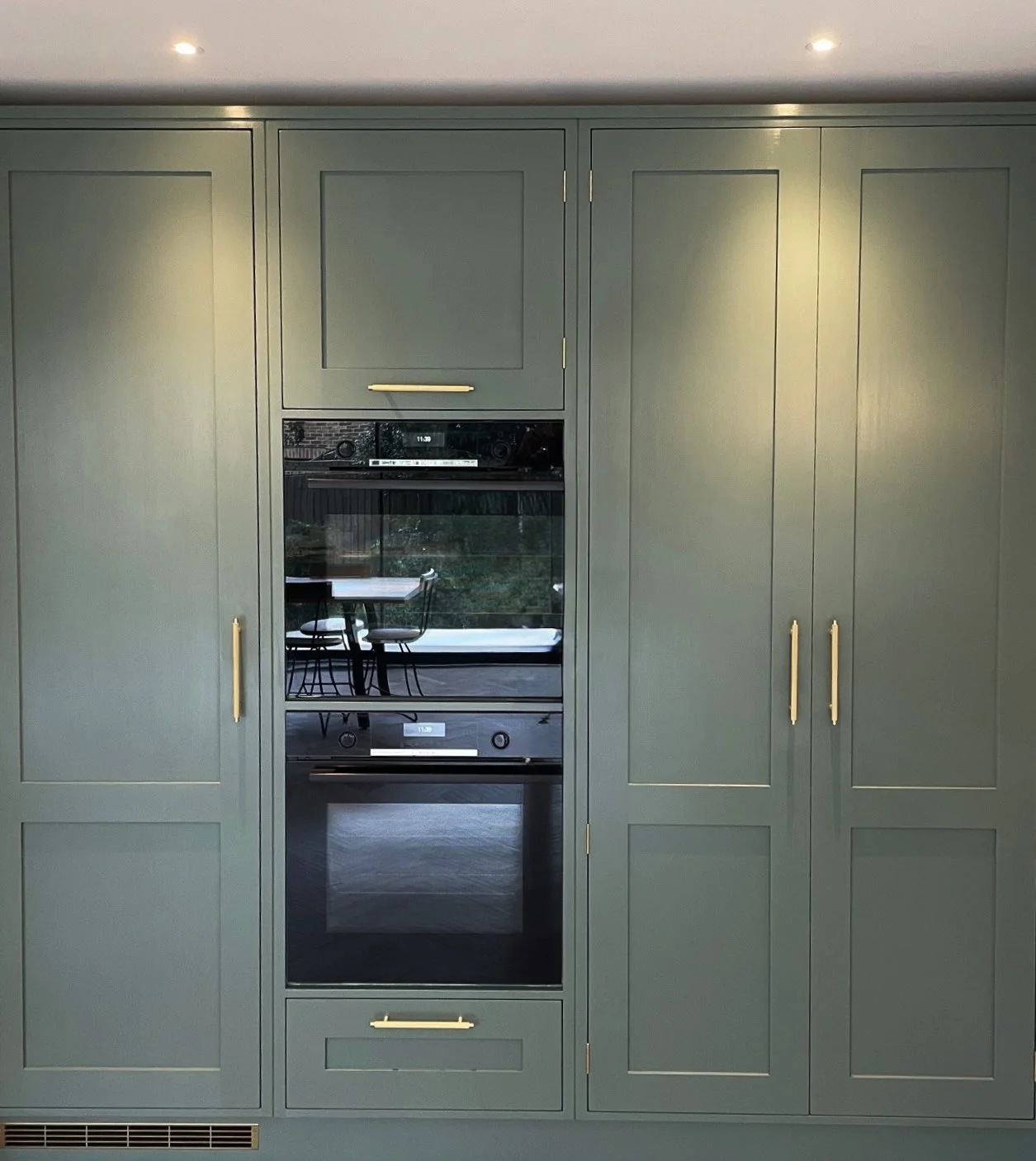 Green kitchen cabinets with built-in oven and microwave, with gold handles, and a window reflecting a table and chairs outside.