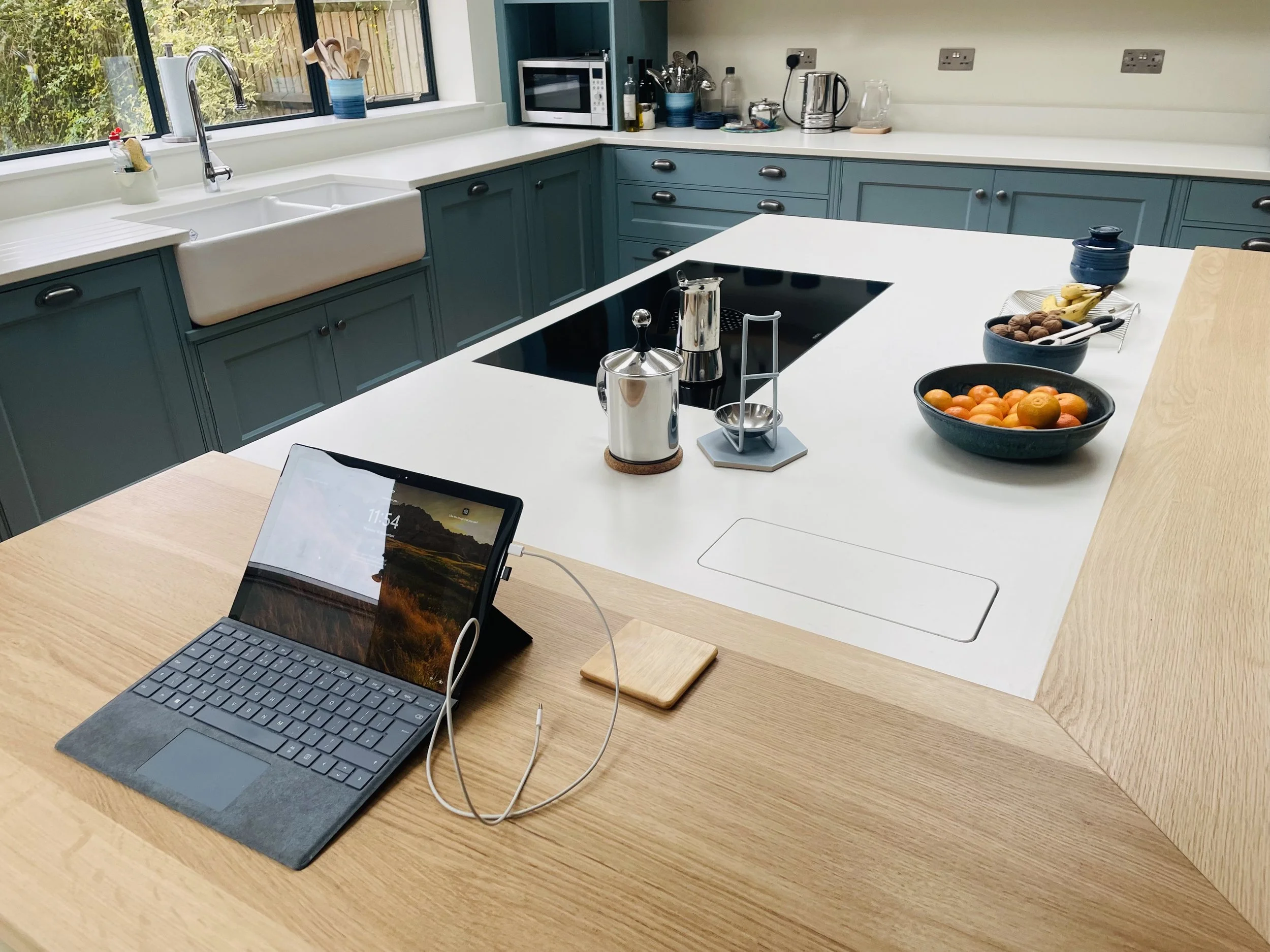 Modern kitchen with teal cabinets, white countertop, farmhouse sink, window with outdoor view, and various kitchen appliances including microwave, kettle, and utensils. On the island, there are bowls of fruit, a coffee or tea kettle, and a tablet on 